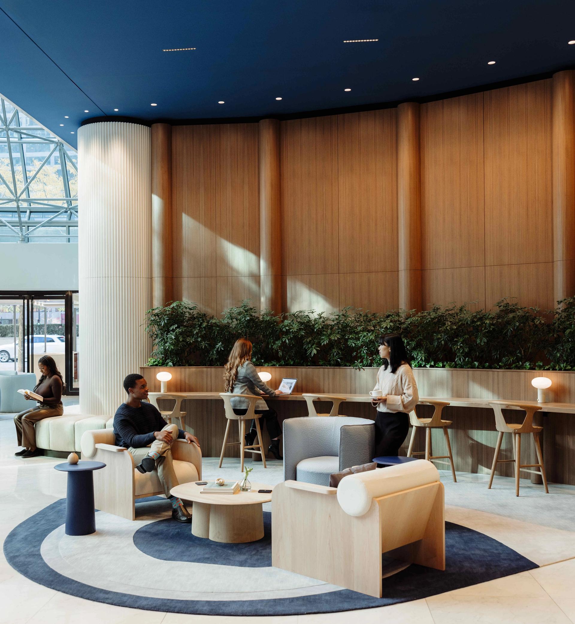 Lobby lounge at 525 West Monroe with modern seating, circular rugs, tall wooden panel walls, built-in planter greenery, and people working on laptops under natural light from the glass atrium ceiling.