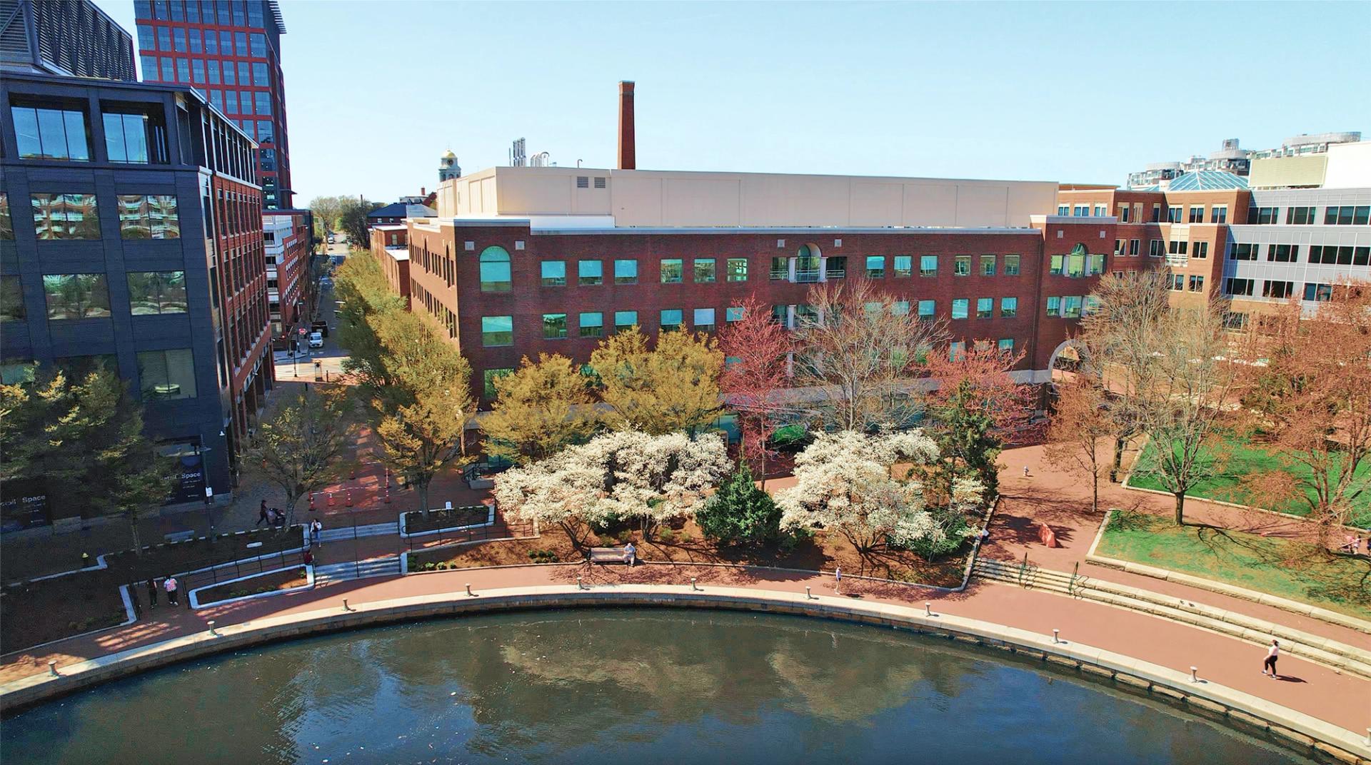 Aerial view of a modern office building with red brick facade, surrounded by trees and a walking path next to a body of water on a clear day.