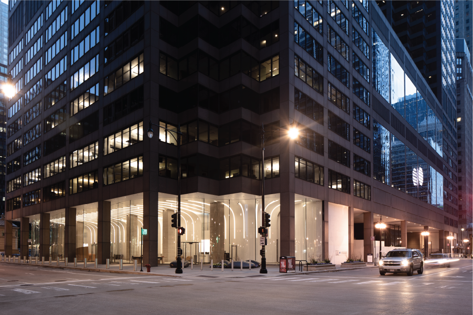 A modern, dark-colored building with reflective windows stands on a bustling city street, illuminated by street lamps and a car driving by, while tall buildings and traffic lights line the street.