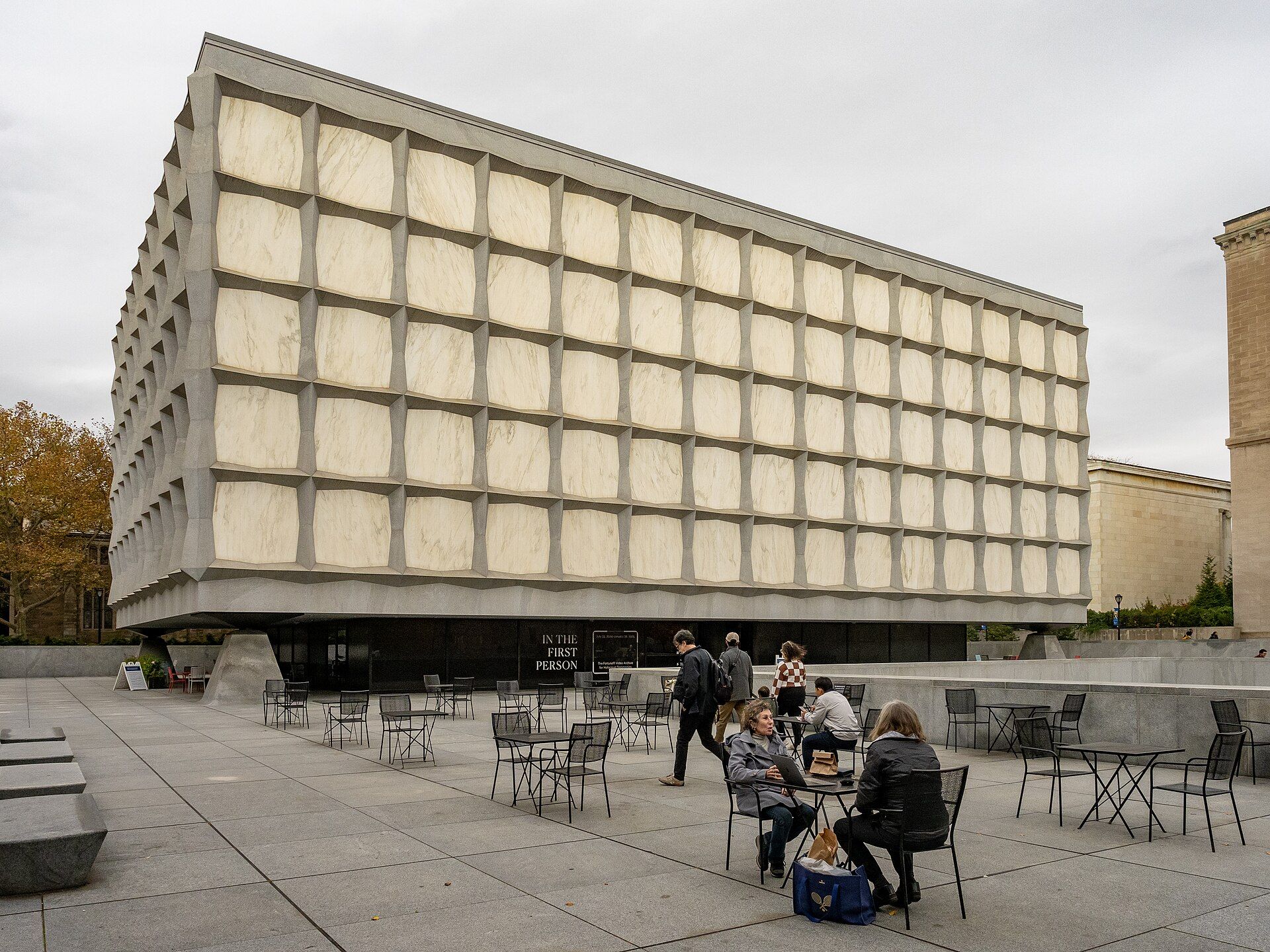 A large, modern building with a square facade and a large glass panel is surrounded by a courtyard with tables and chairs, where people are enjoying their time.