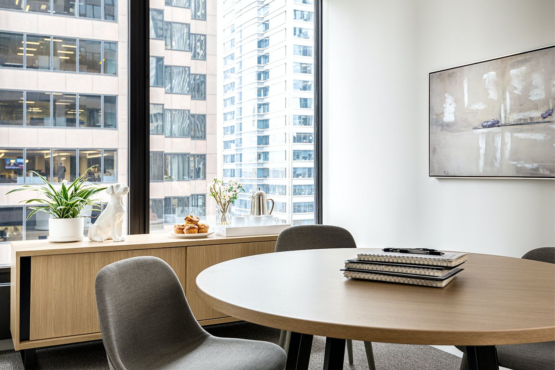 A modern office with a wooden desk, white chair, and a large window overlooking a cityscape, featuring a painting, potted plant, and a vase of flowers.