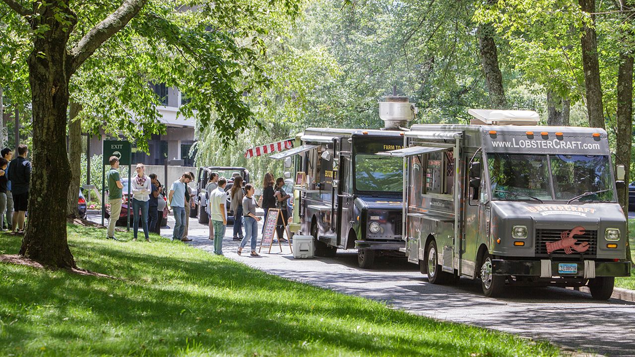 A bustling park scene features a line of food trucks parked on a sidewalk, surrounded by lush greenery and people enjoying their meals.