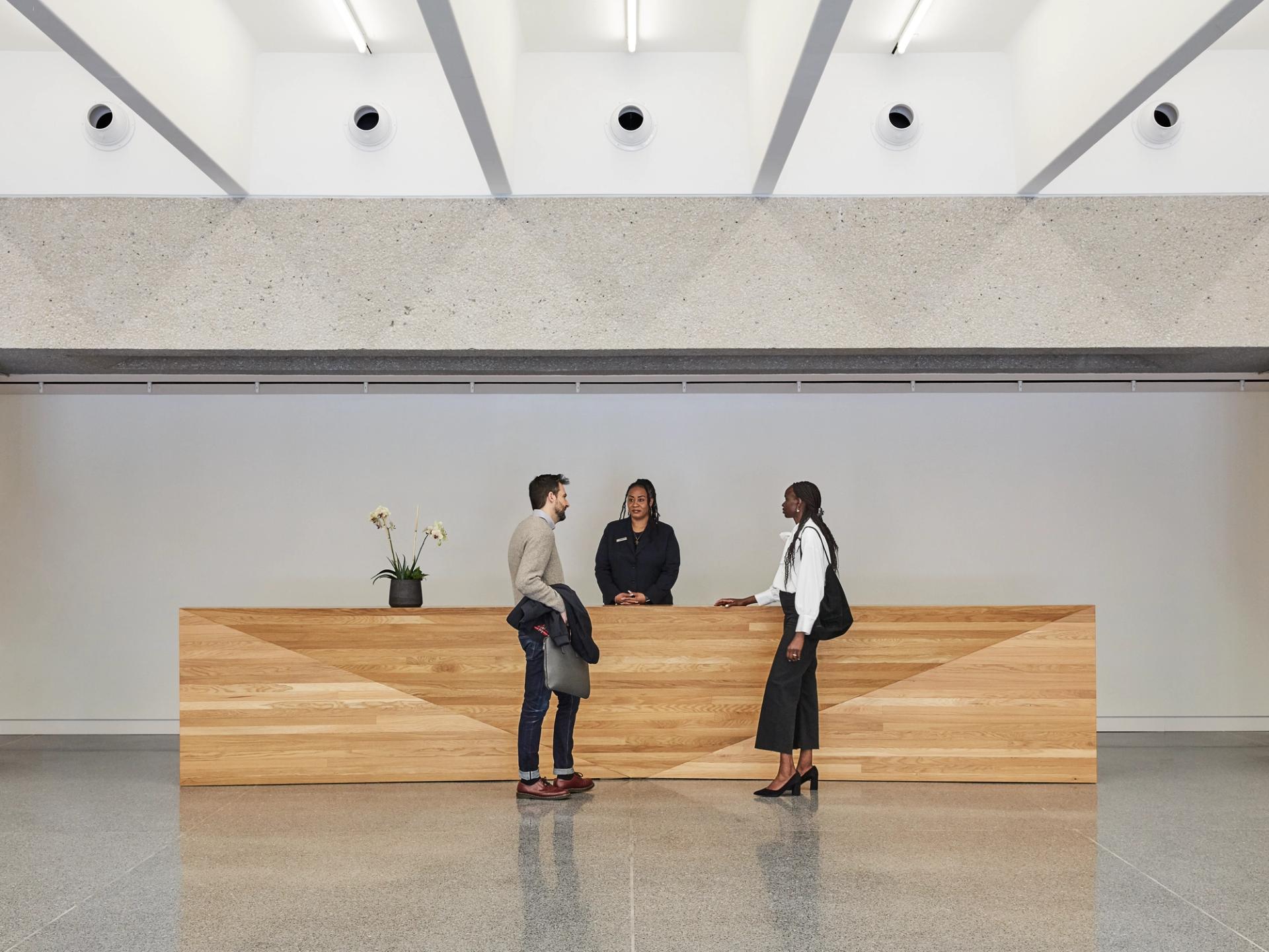 A modern, spacious lobby features a wooden reception desk with a potted plant, two women conversing behind it, and a large window providing natural light. 