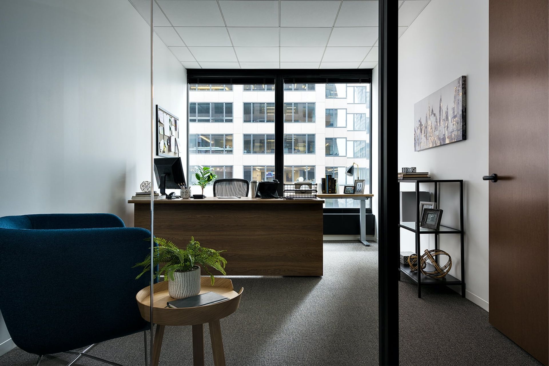 A modern office with a desk, chair, and plant, a wooden door, and a large window with a view of a building.