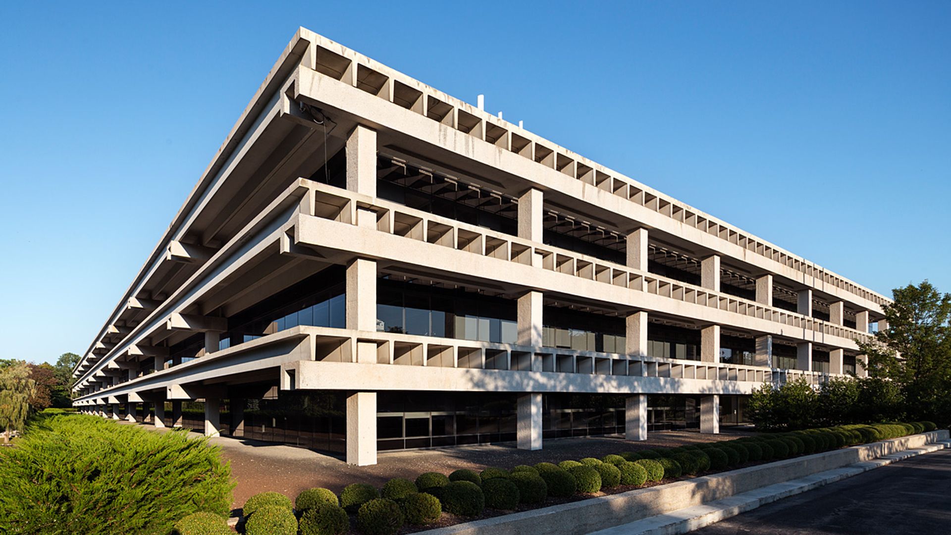 A modern, multi-level building with a concrete facade and numerous windows stands in a lush green landscape, surrounded by trees and shrubs, with a person walking nearby.