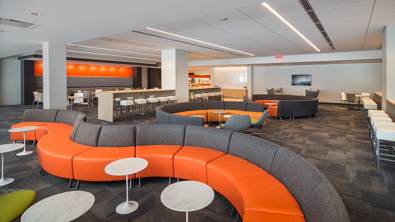 The image shows a modern, spacious office with a large, curved seating area, a bar, and a bright, well-lit atmosphere.