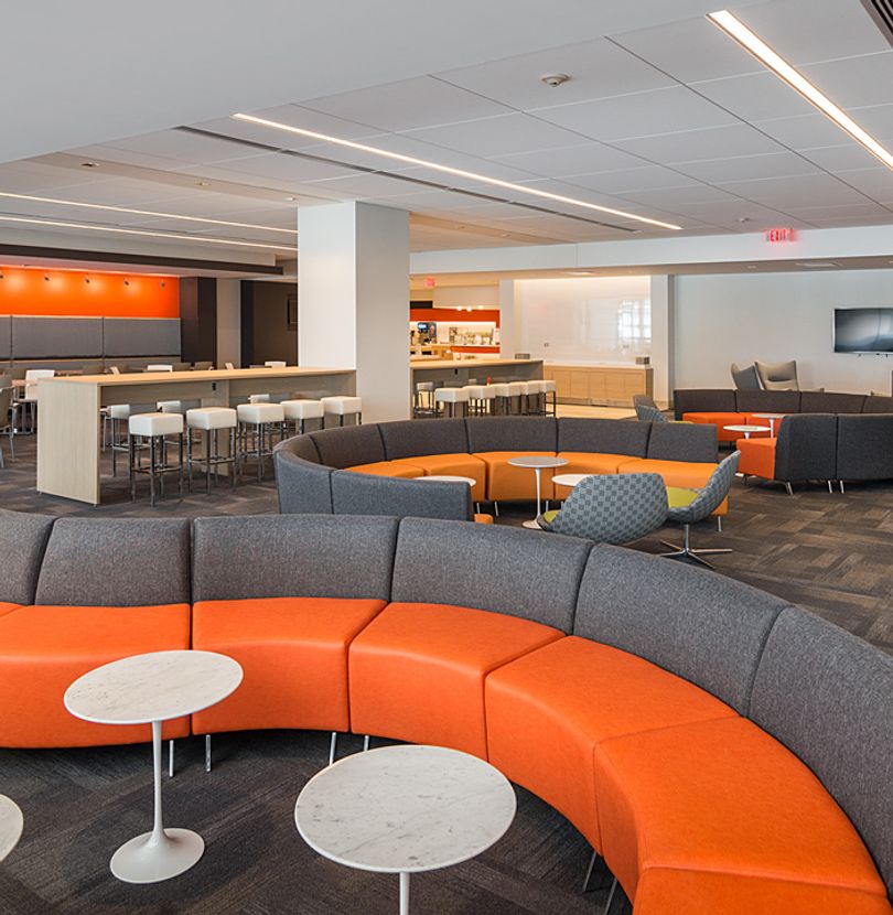 The image shows a modern, spacious office with a large, curved seating area, a bar, and a bright, well-lit atmosphere.