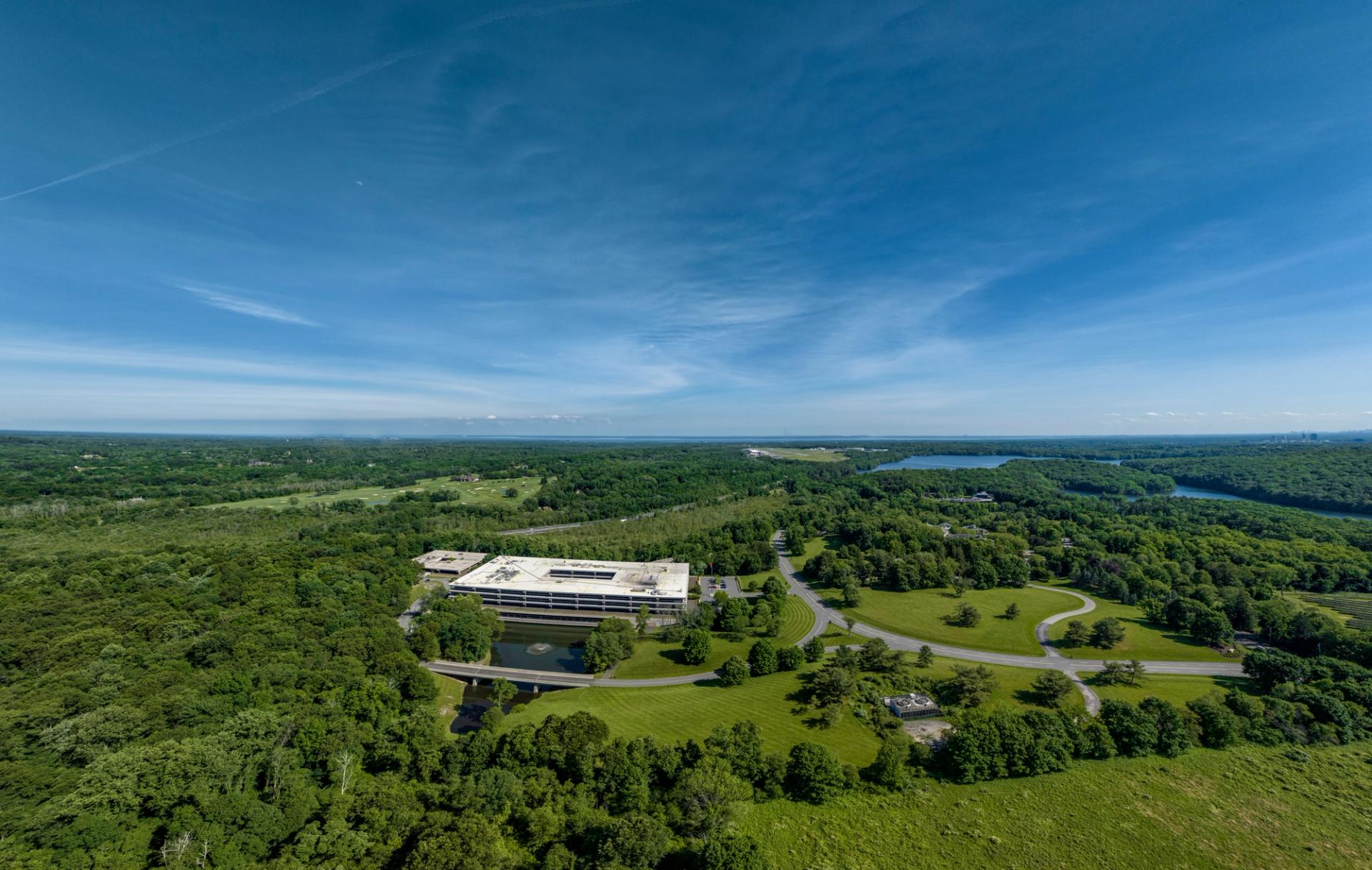 An aerial view of a large white building surrounded by greenery, with a road and a lake in the background, showcasing a harmonious blend of urban and natural elements.