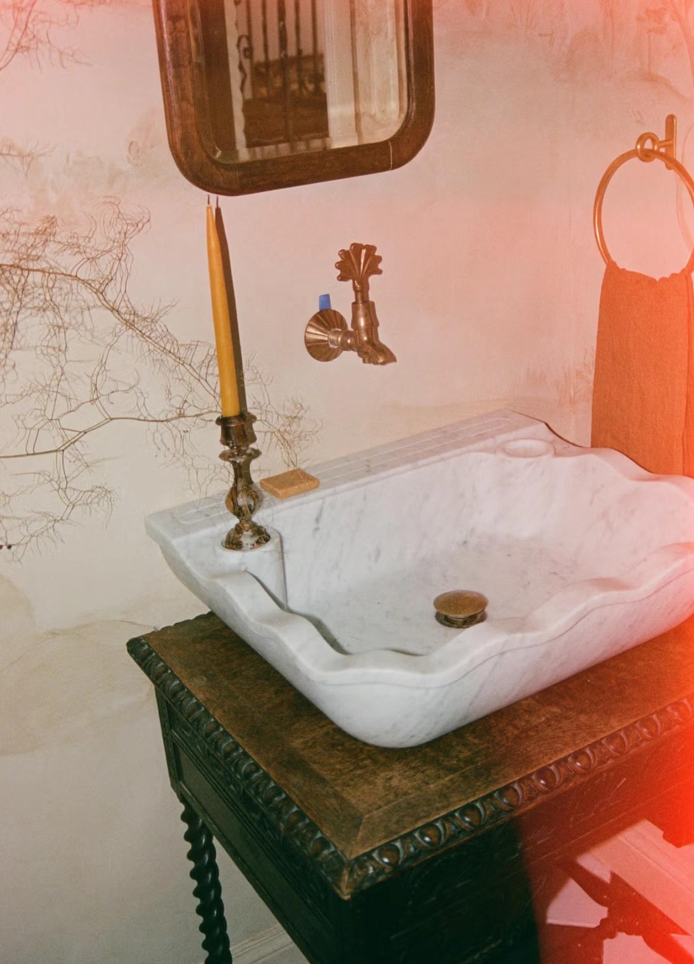 A vintage marble sink rests on an ornate wooden table, with a candle holder and a towel nearby against a textured wall.