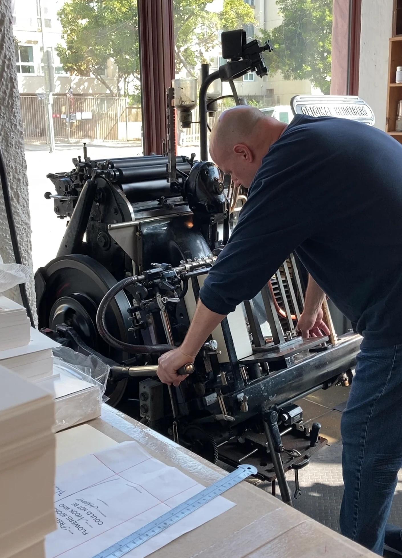 A person adjusts an old printing press, surrounded by stacks of paper and sunlight streaming through large windows.