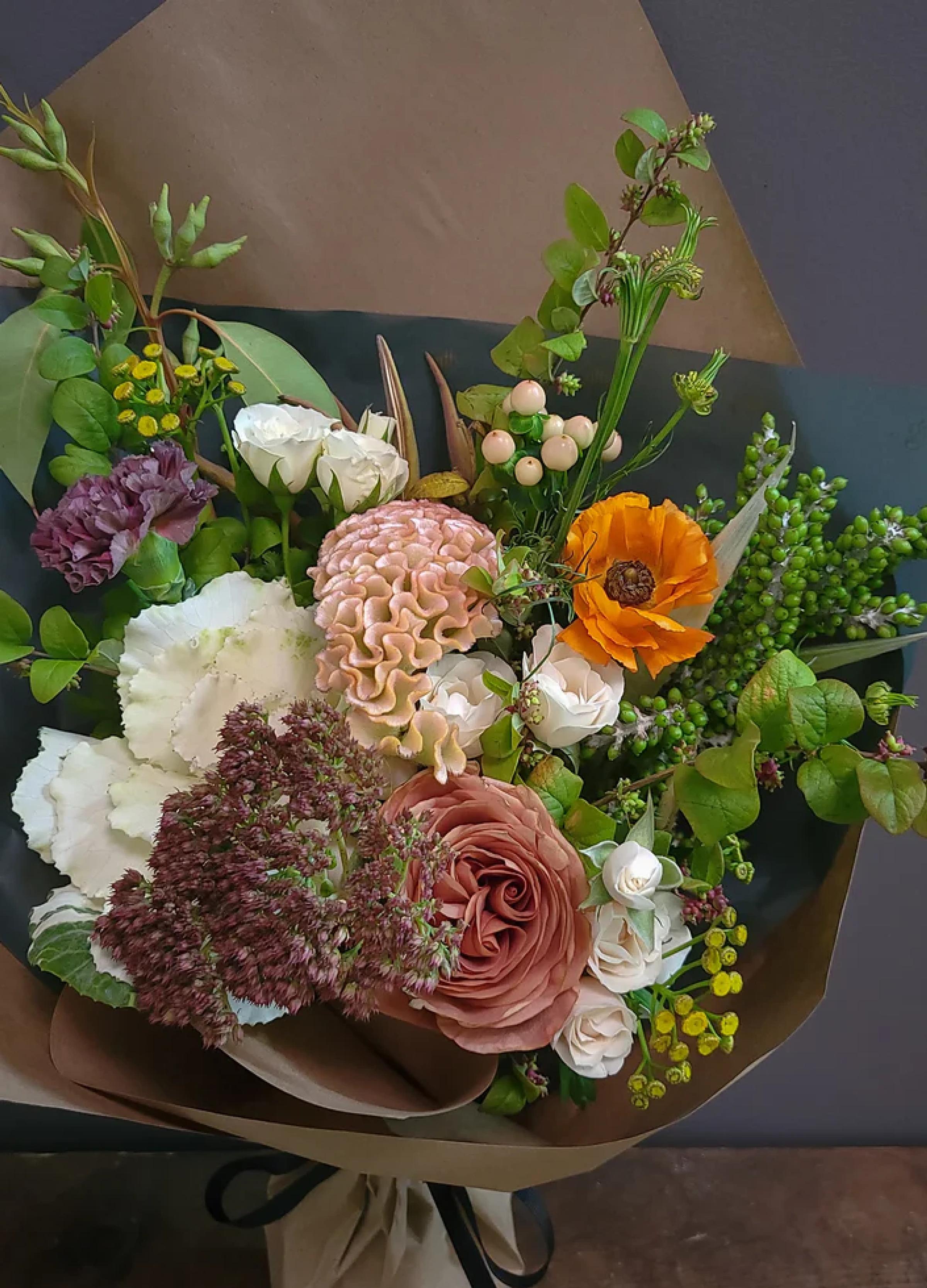 A vibrant bouquet featuring roses, peonies, and greenery, wrapped in brown paper with a black ribbon bow.