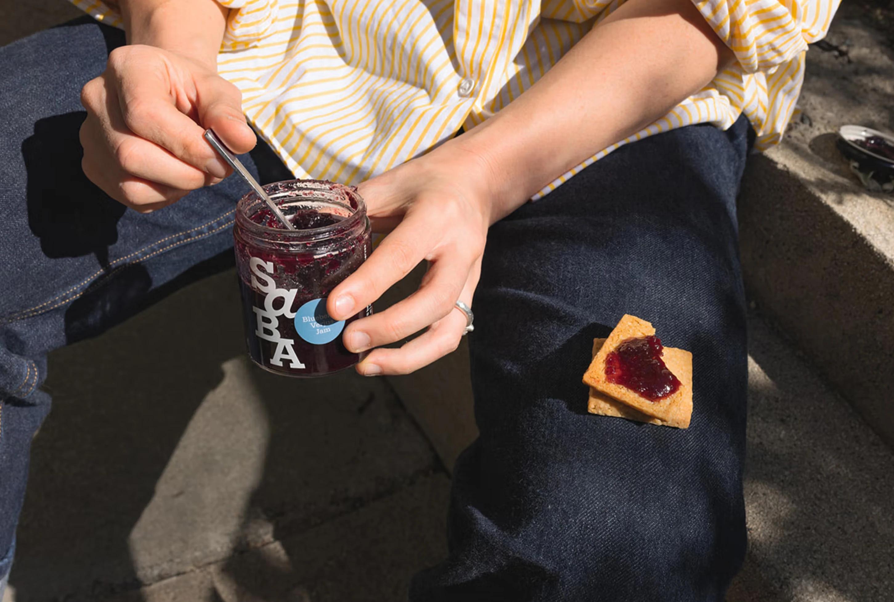 A hand holding a jar of blueberry jam, with a spoon and a cracker topped with jam on a lap. Sunlight casts soft shadows.