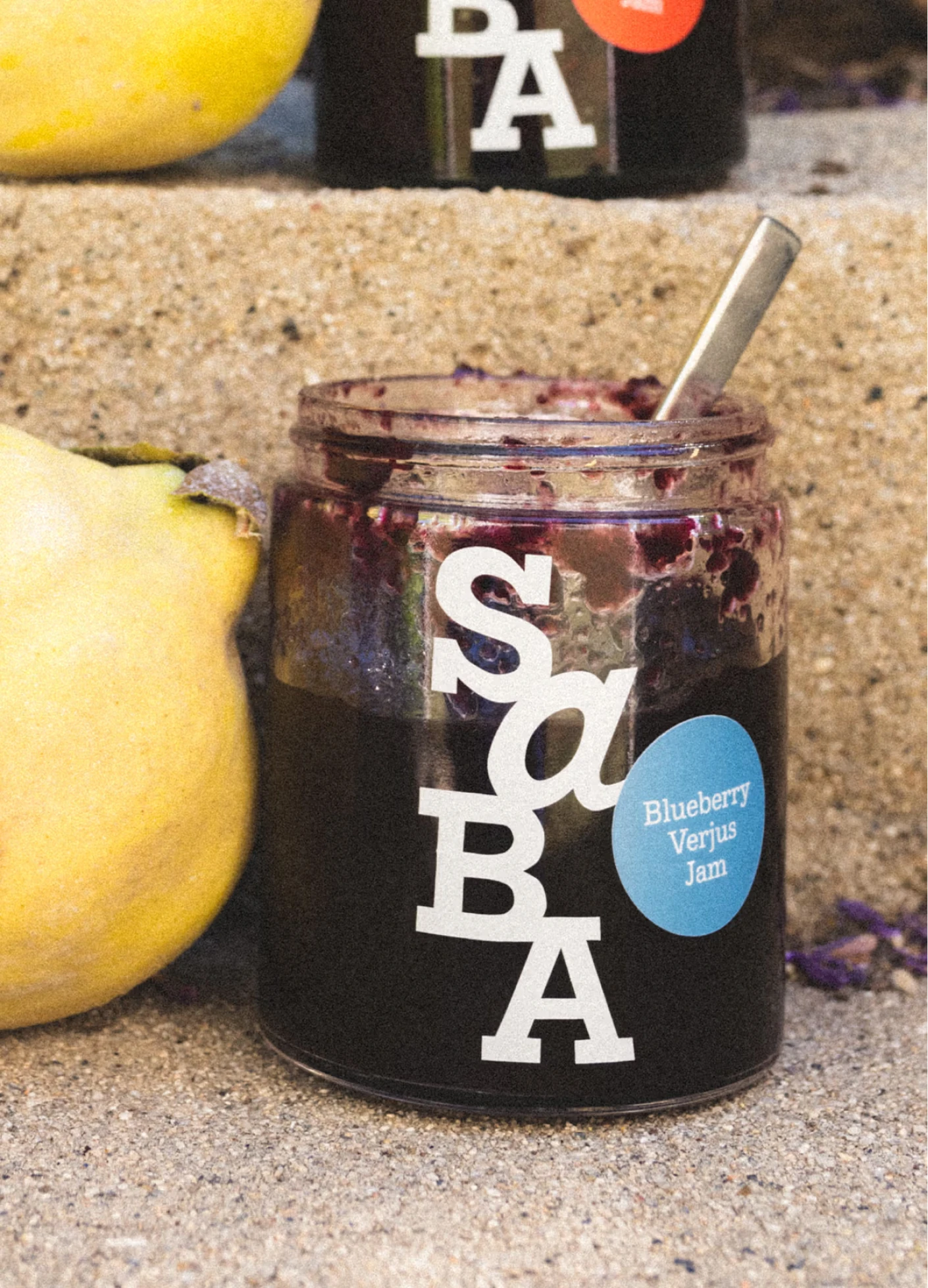 A jar of blueberry verjus jam with a spoon, next to a yellow quince on a textured surface.