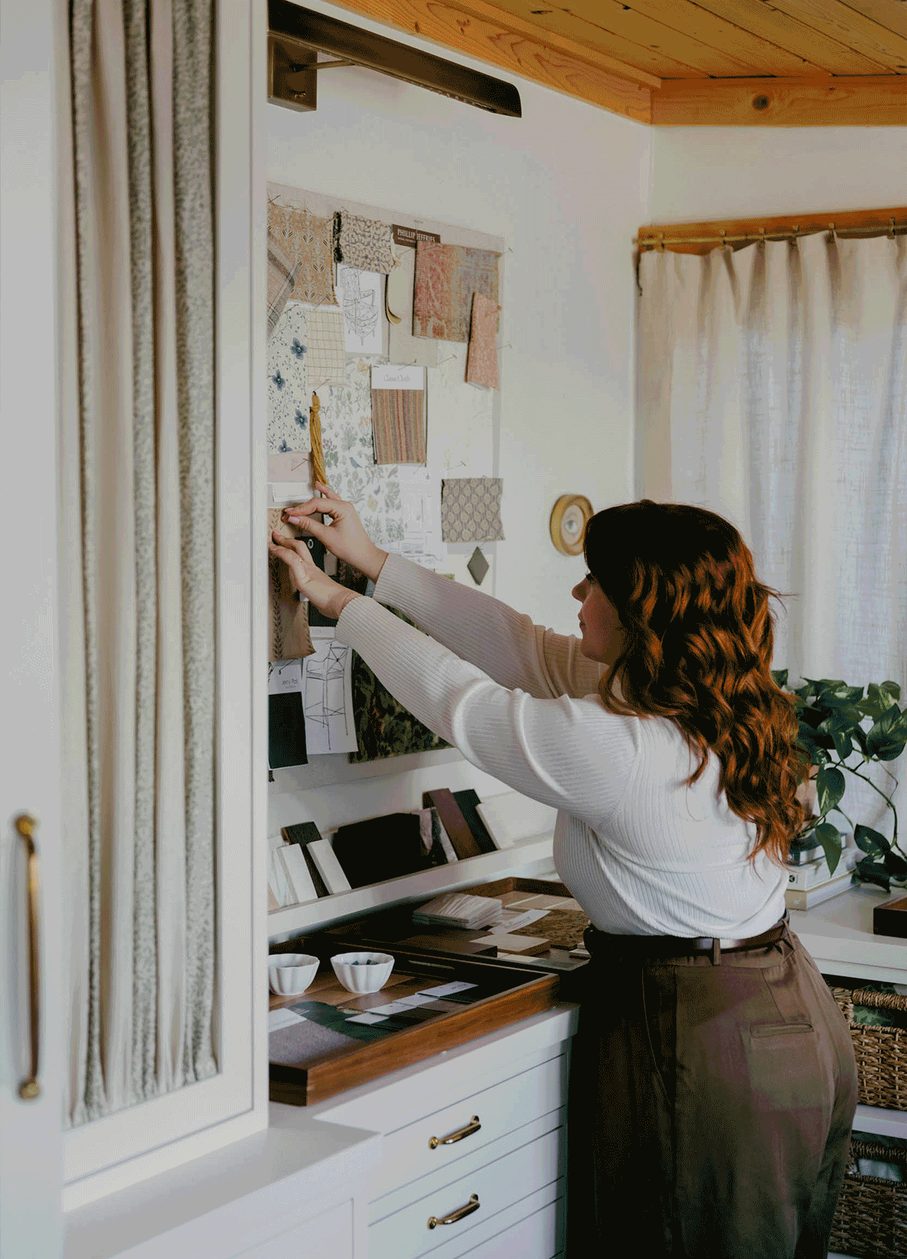 A woman with wavy hair pins fabric swatches and sketches on a mood board in a bright, stylish workspace.