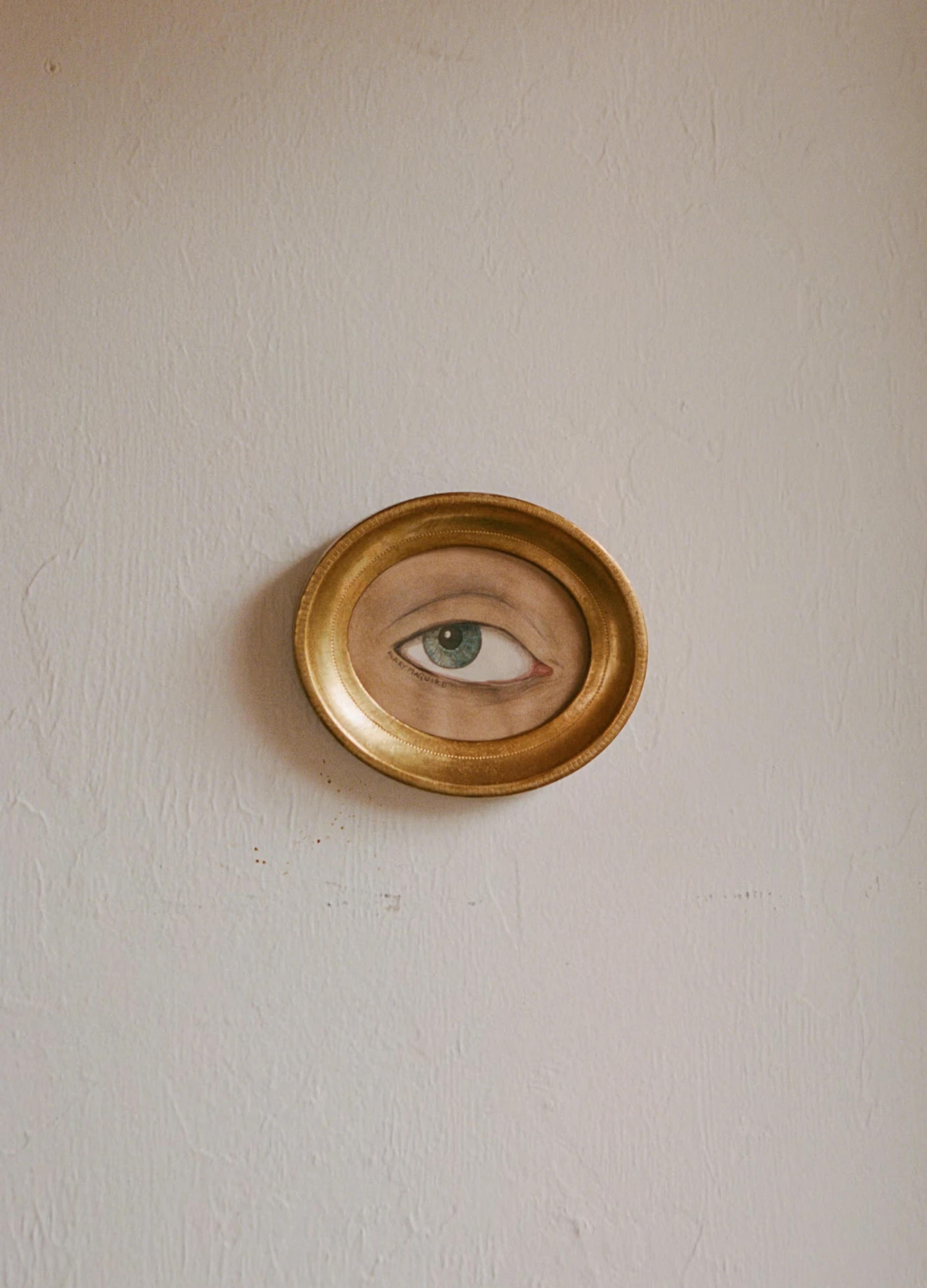 A painted eye encircled by a gold frame, mounted on a textured white wall.