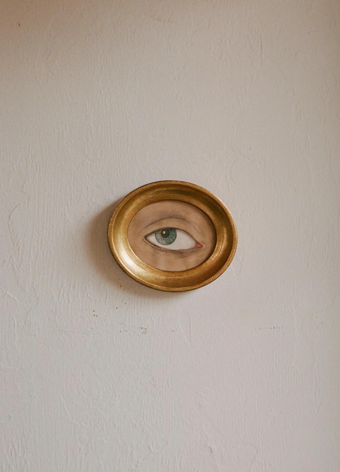A painted eye encircled by a gold frame, mounted on a textured white wall.