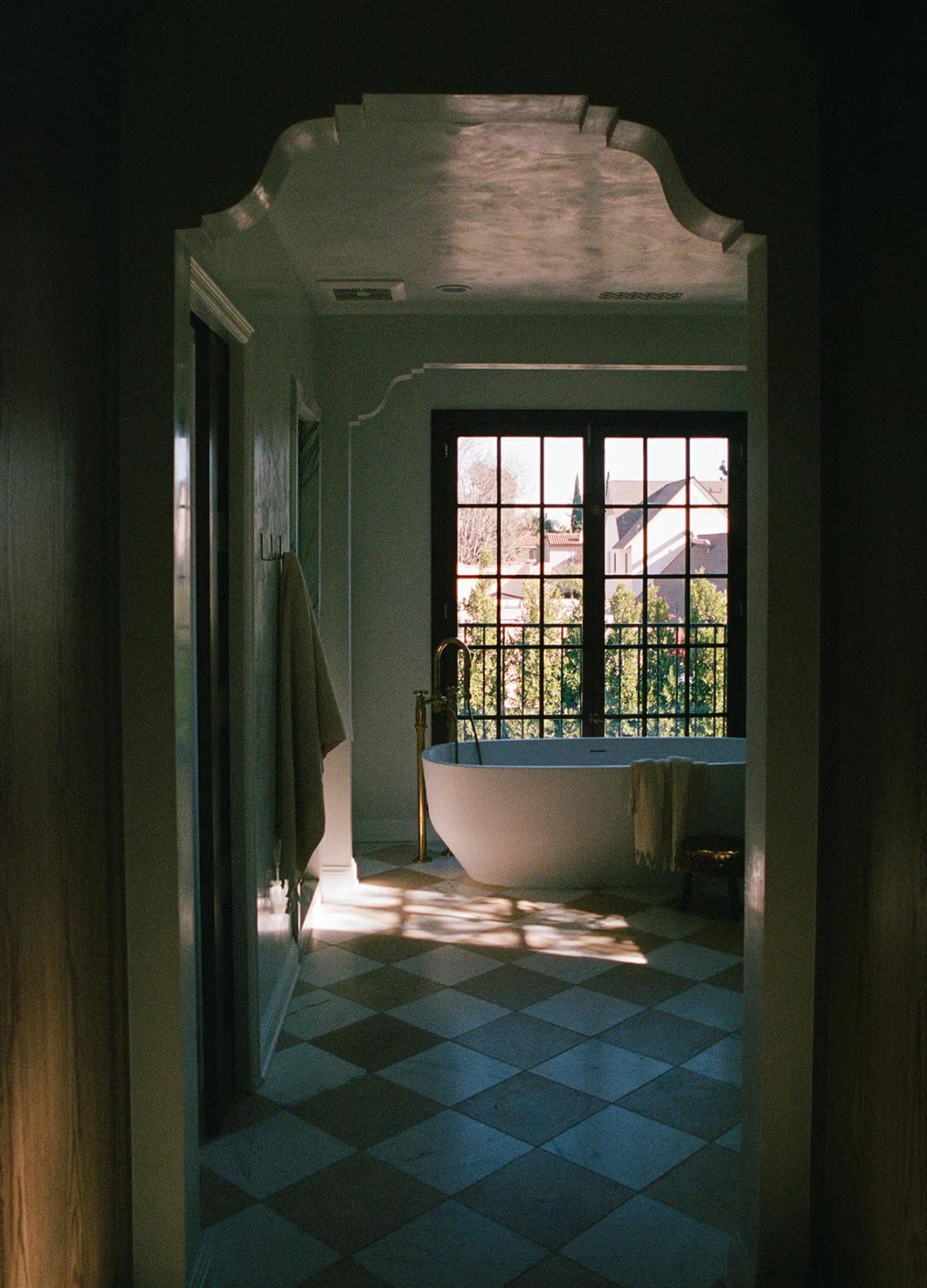 Bright, stylish bathroom featuring a freestanding tub, large window, and checkered stone floor, with soft shadows playing ...