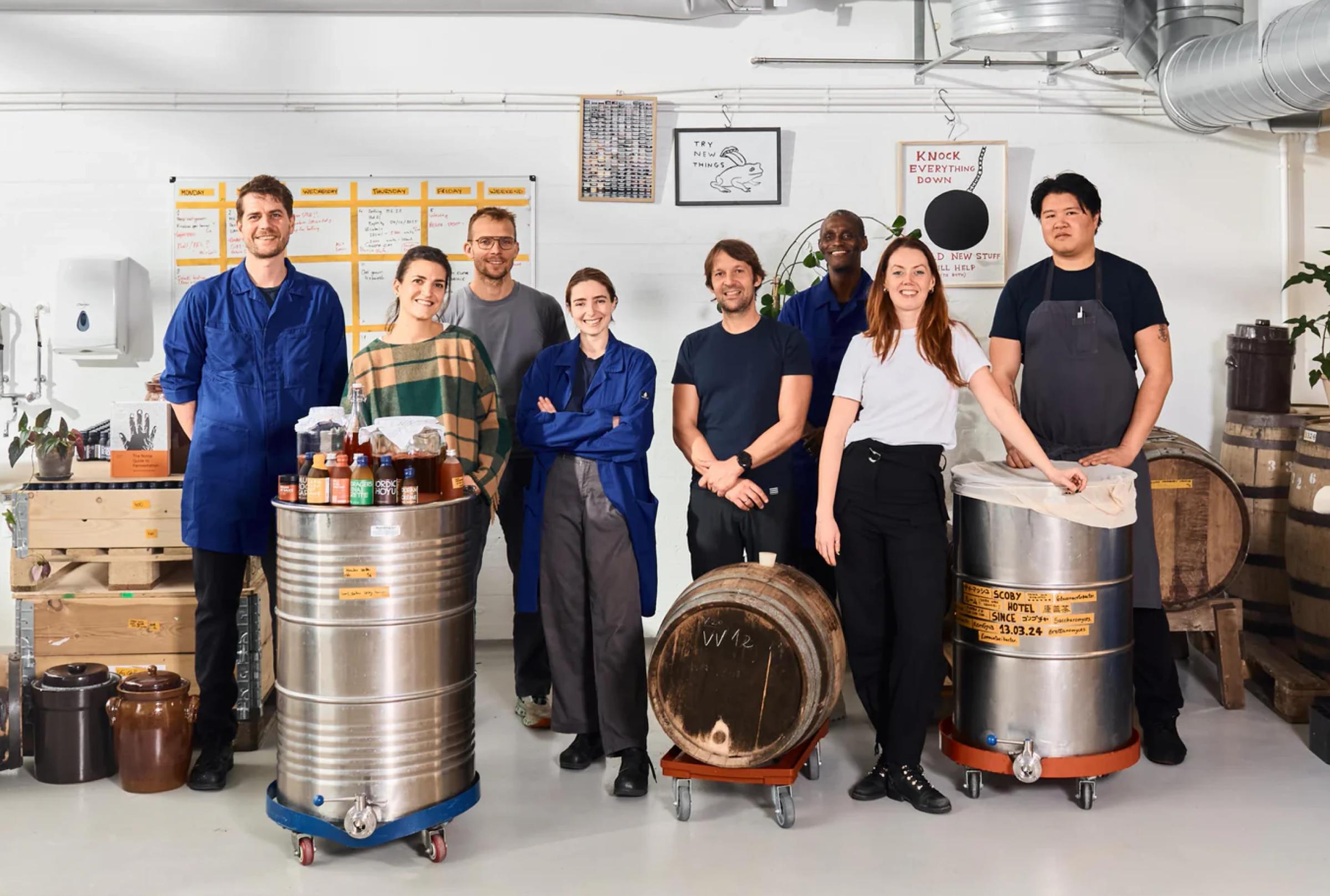 A diverse group of eight people stands in a bright workspace, surrounded by barrels and brewing equipment.
