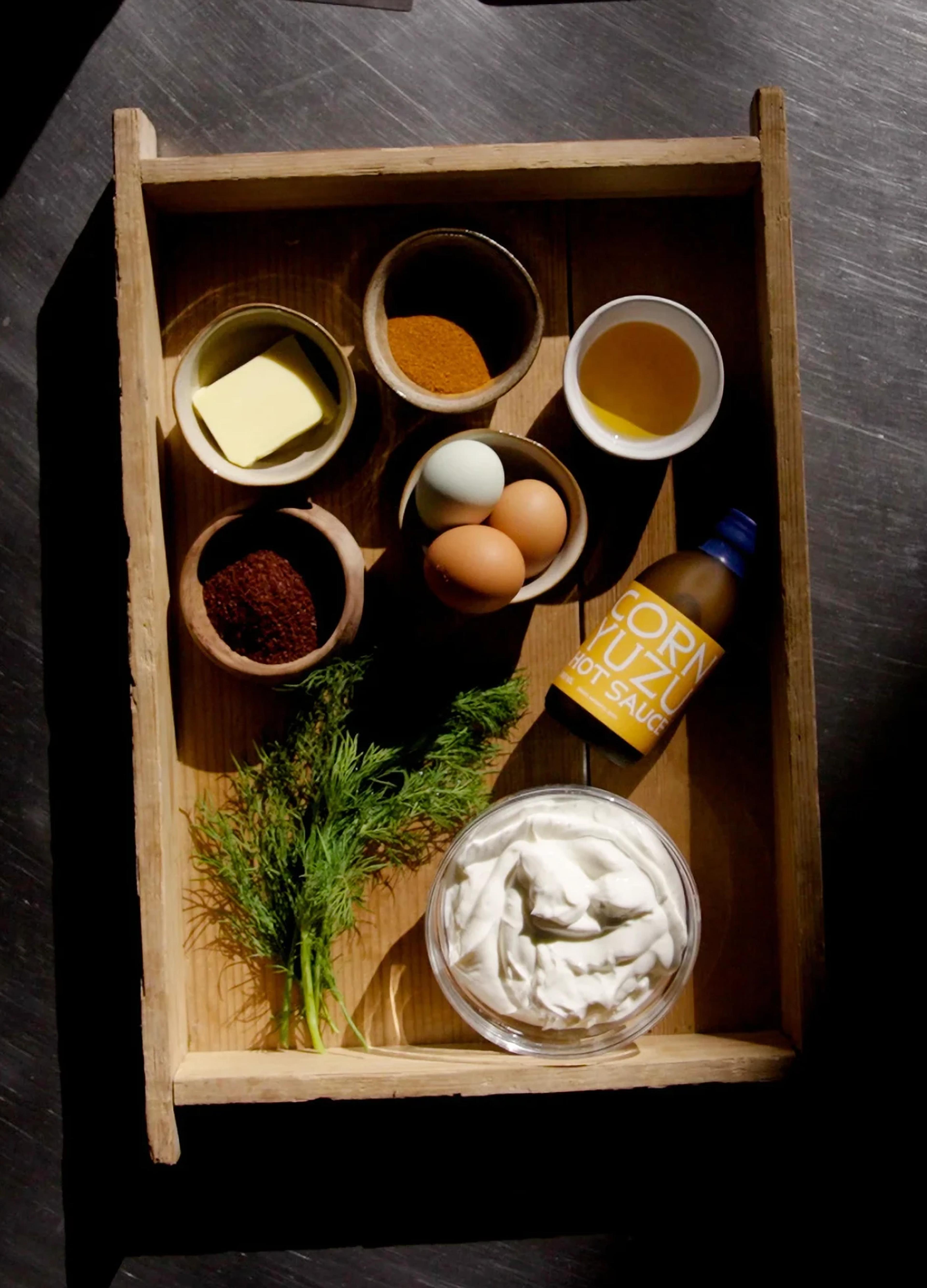 A wooden tray holds butter, spices, eggs, olive oil, hot sauce, dill, and a bowl of creamy dip.