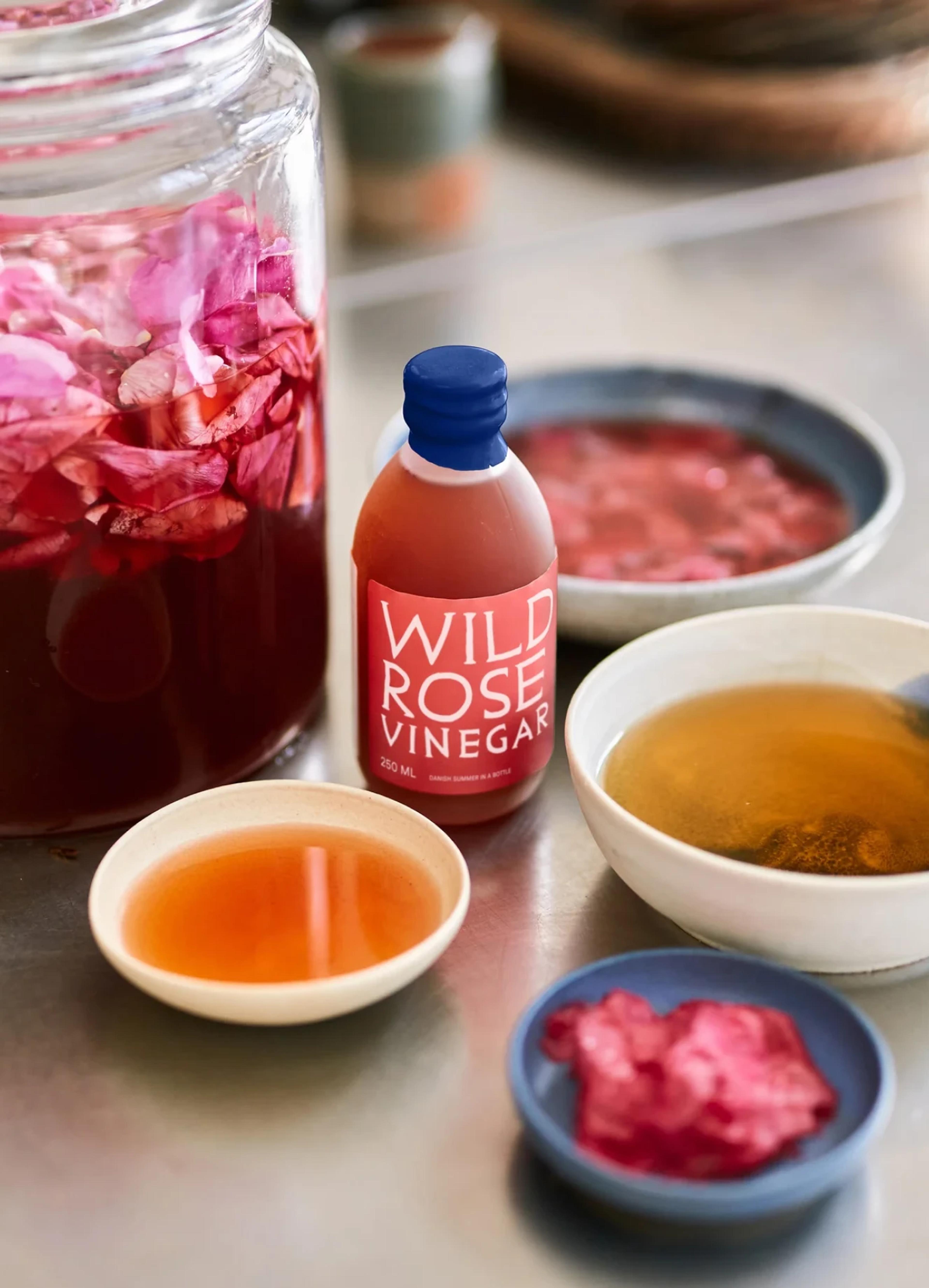 A bottle of Wild Rose Vinegar surrounded by bowls of liquid and rose petals, on a bright countertop.