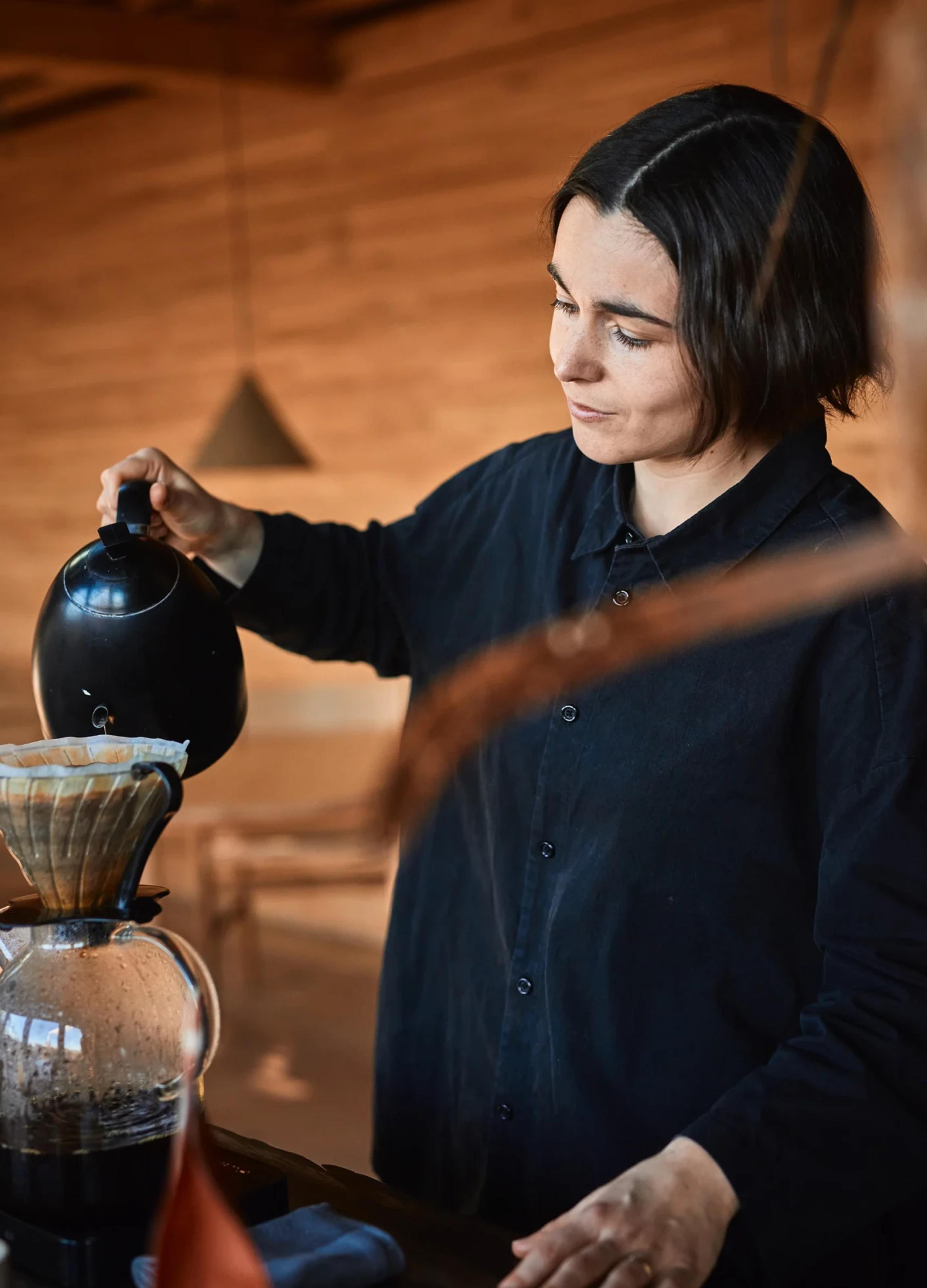 A woman pours hot water from a kettle into a coffee dripper, preparing coffee in a cozy, wooden interior.