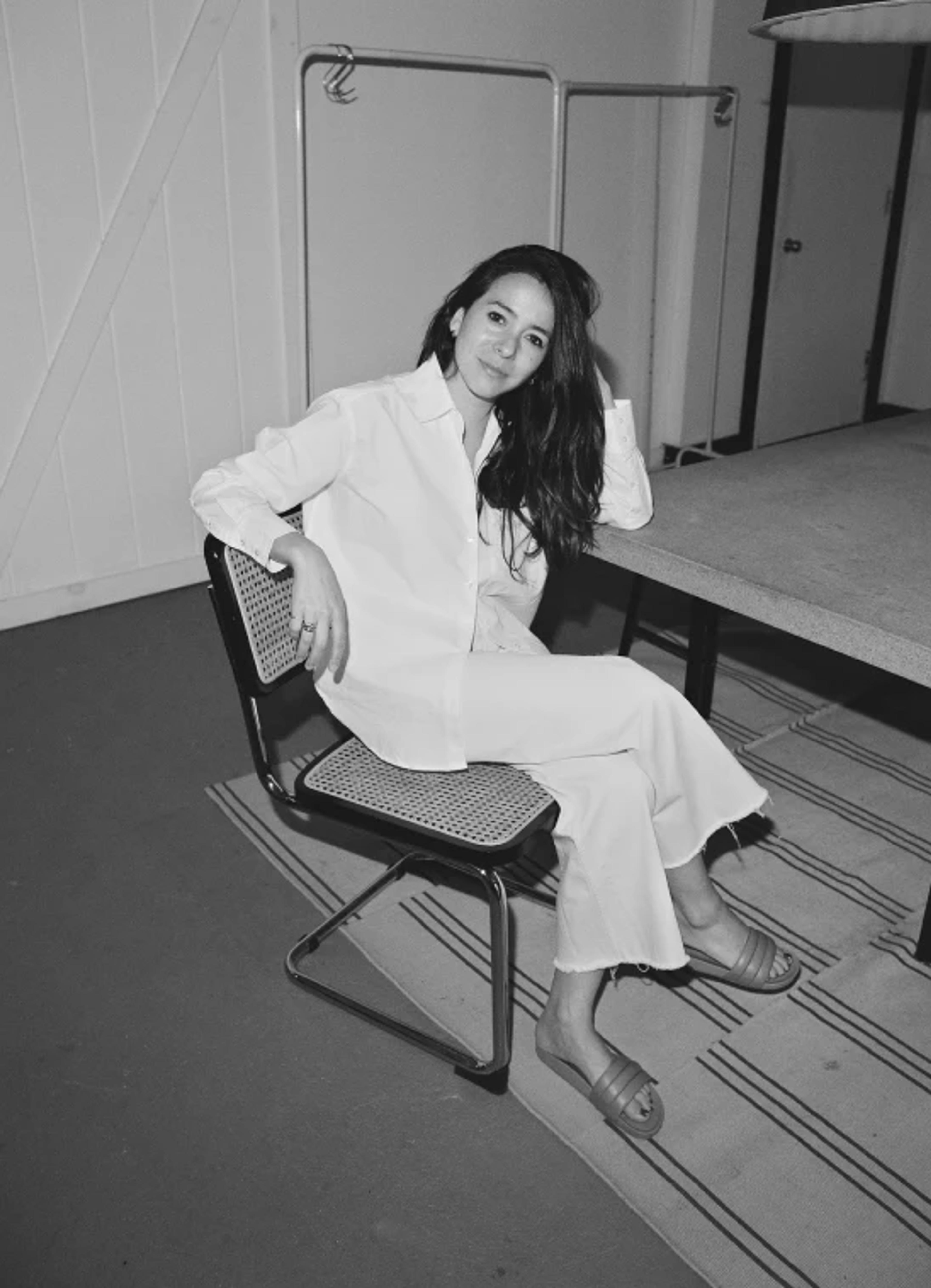 A woman in a white outfit sits casually on a chair, relaxed in a minimalist room with a table and clothing rack.
