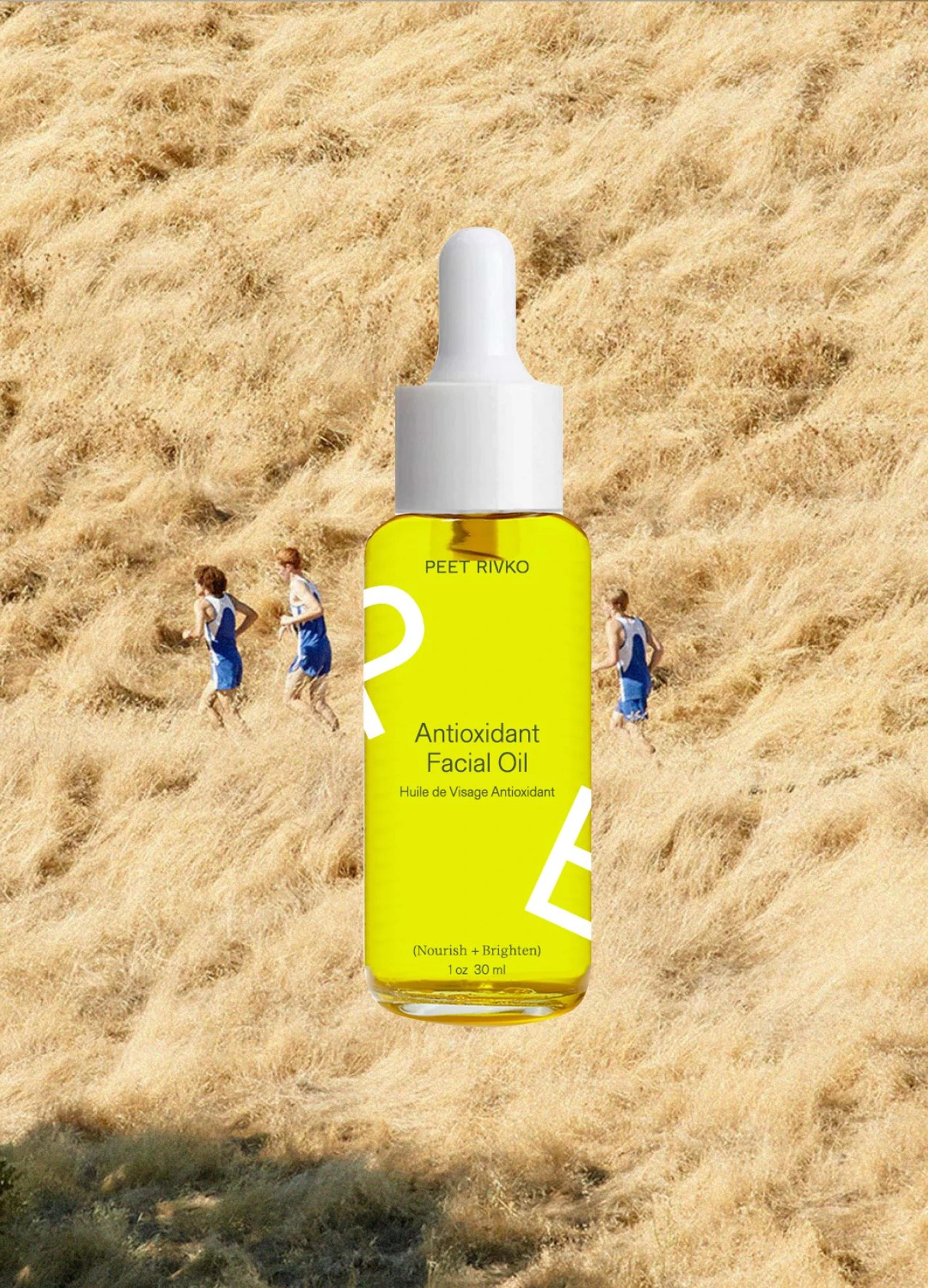 Bright yellow bottle of Antioxidant Facial Oil in front of two people running on a grassy hillside.
