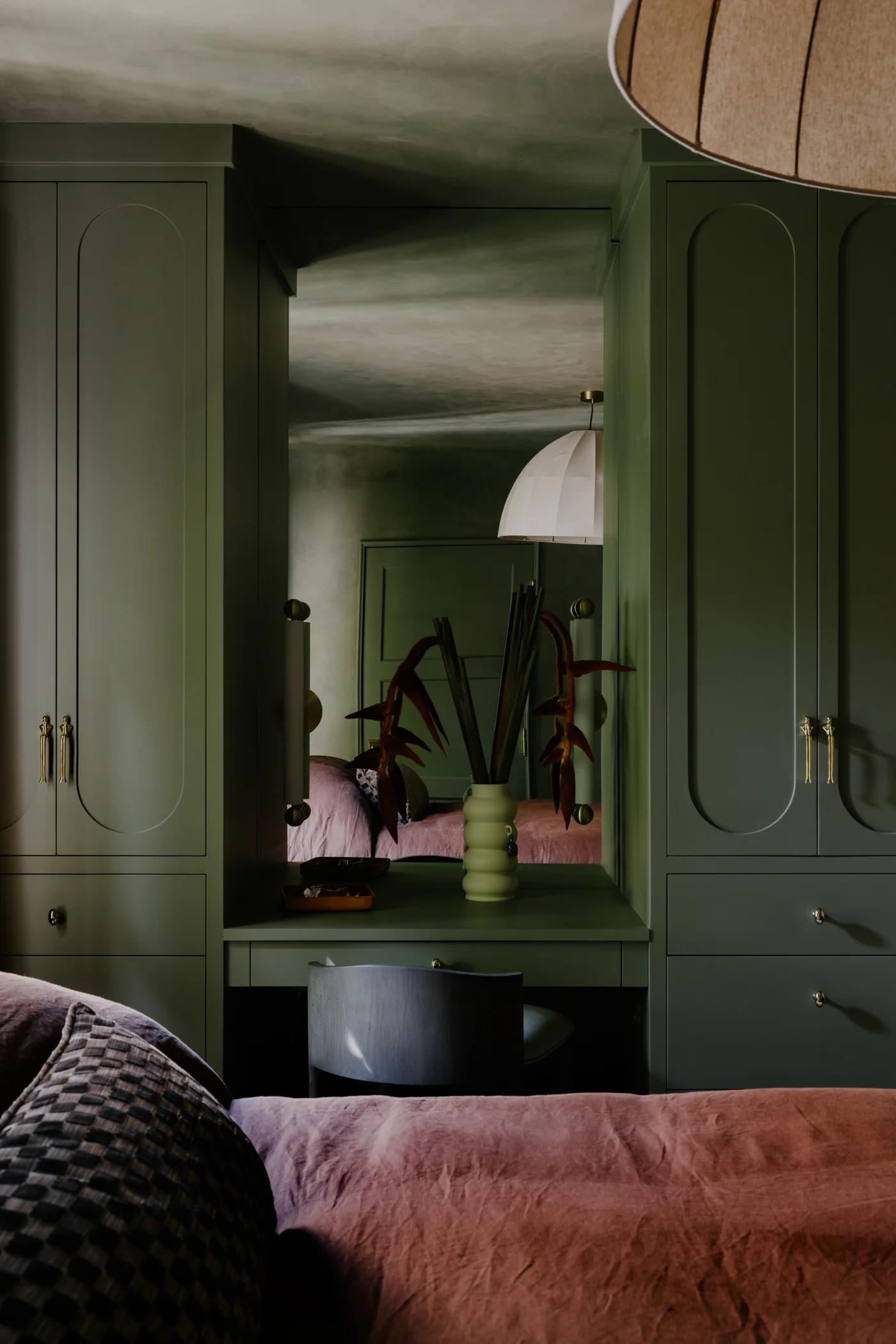 A cozy room featuring green cabinetry, a mirror, a decorative vase with plants, and soft pink bedding.