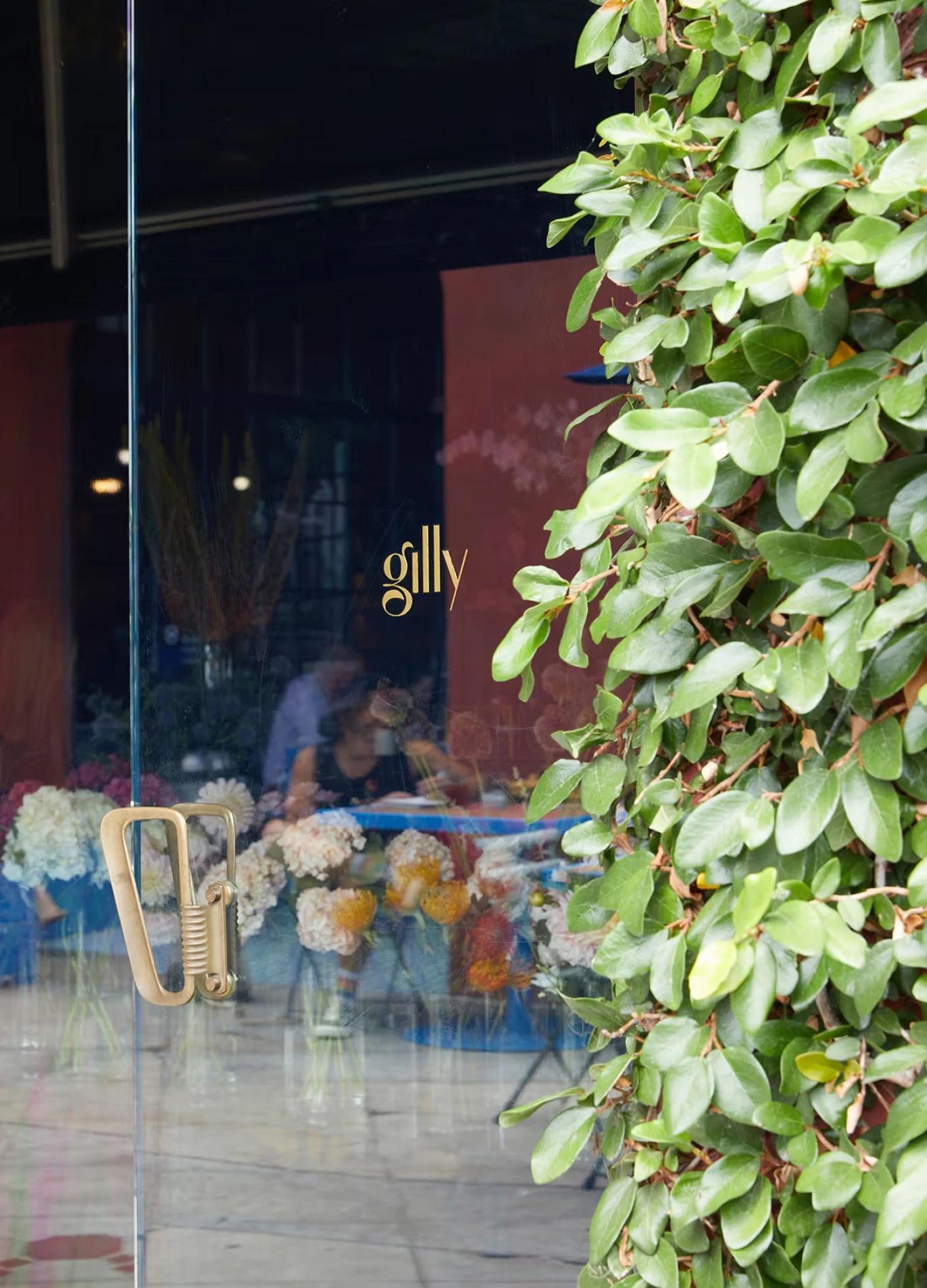 Glass door with gold lettering "gilly," surrounded by green foliage and floral arrangements visible inside.