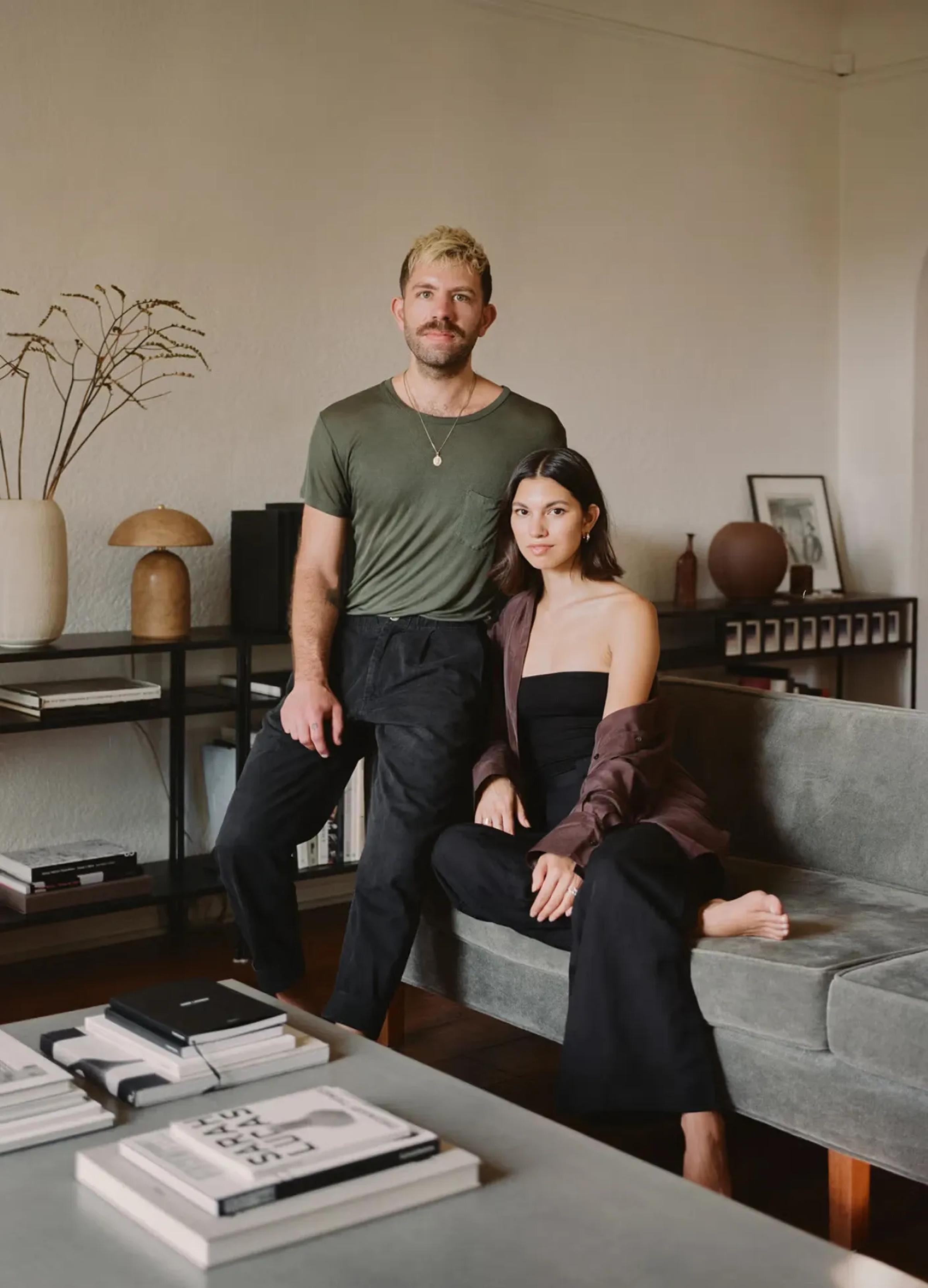 A man and woman pose on a couch in a stylish living room, surrounded by books and minimalist decor.