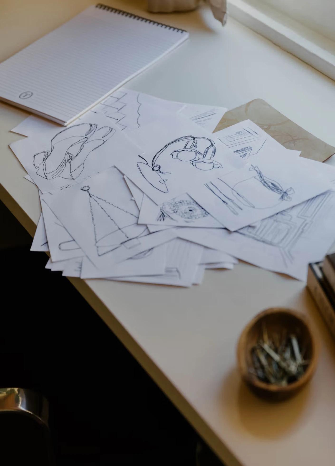 A stack of sketches and design notes scattered on a desk, next to a bowl of small metal fasteners and a notepad.