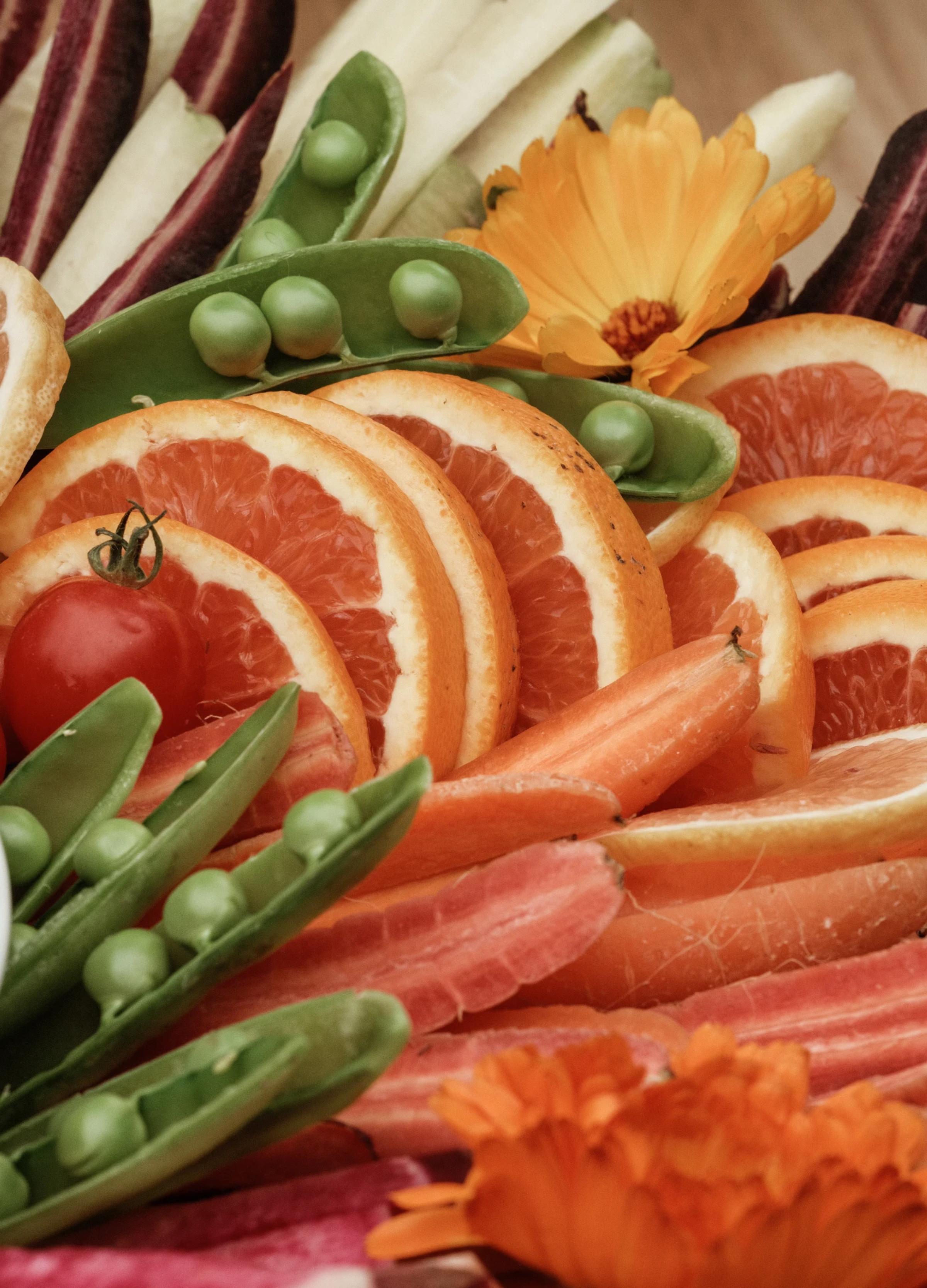 Vibrant assortment of orange slices, cherry tomatoes, carrots, peas, and colorful flowers arranged artfully.