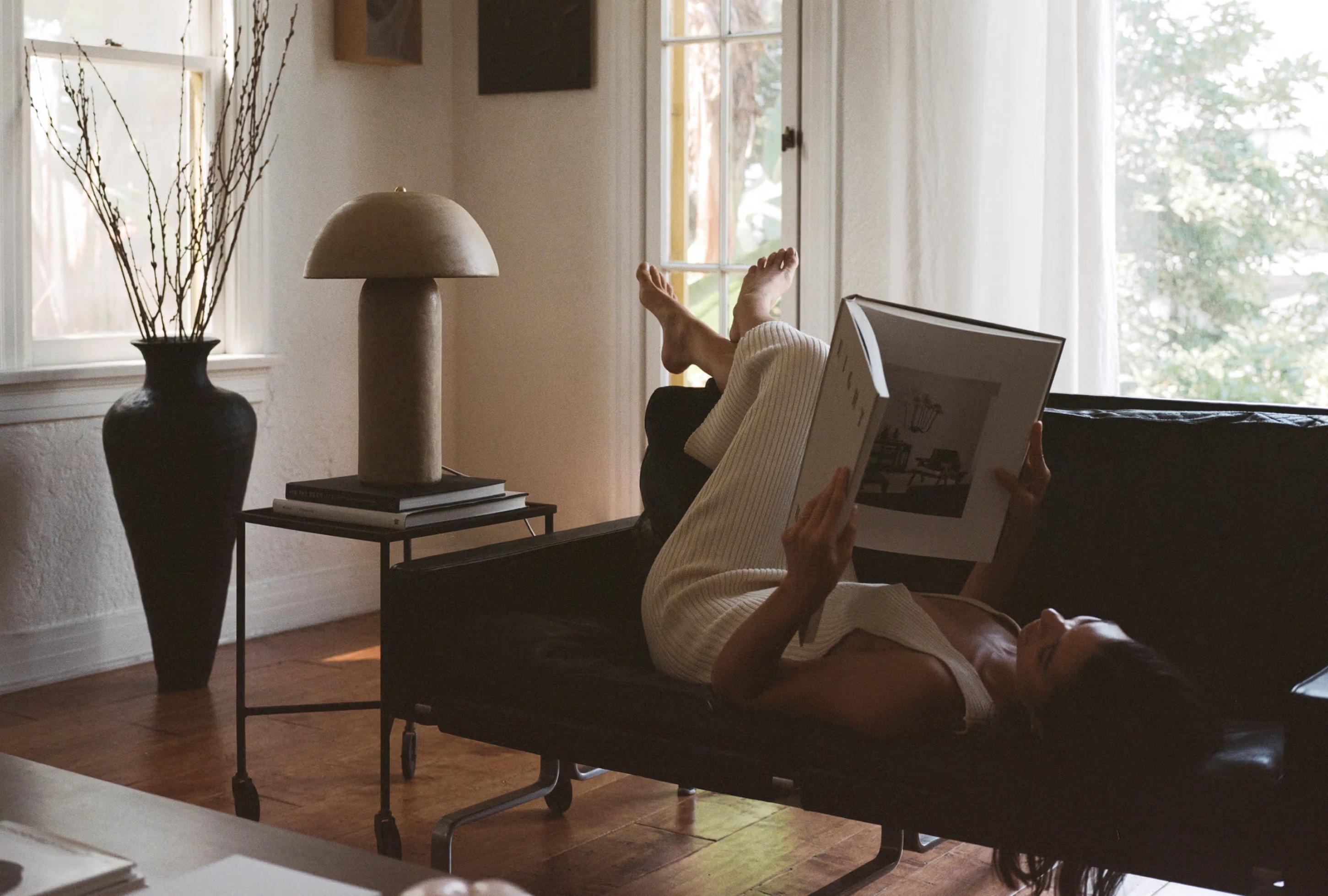 A woman relaxes on a black couch, reading a book, with a stylish lamp and a vase of branches in a well-lit room.