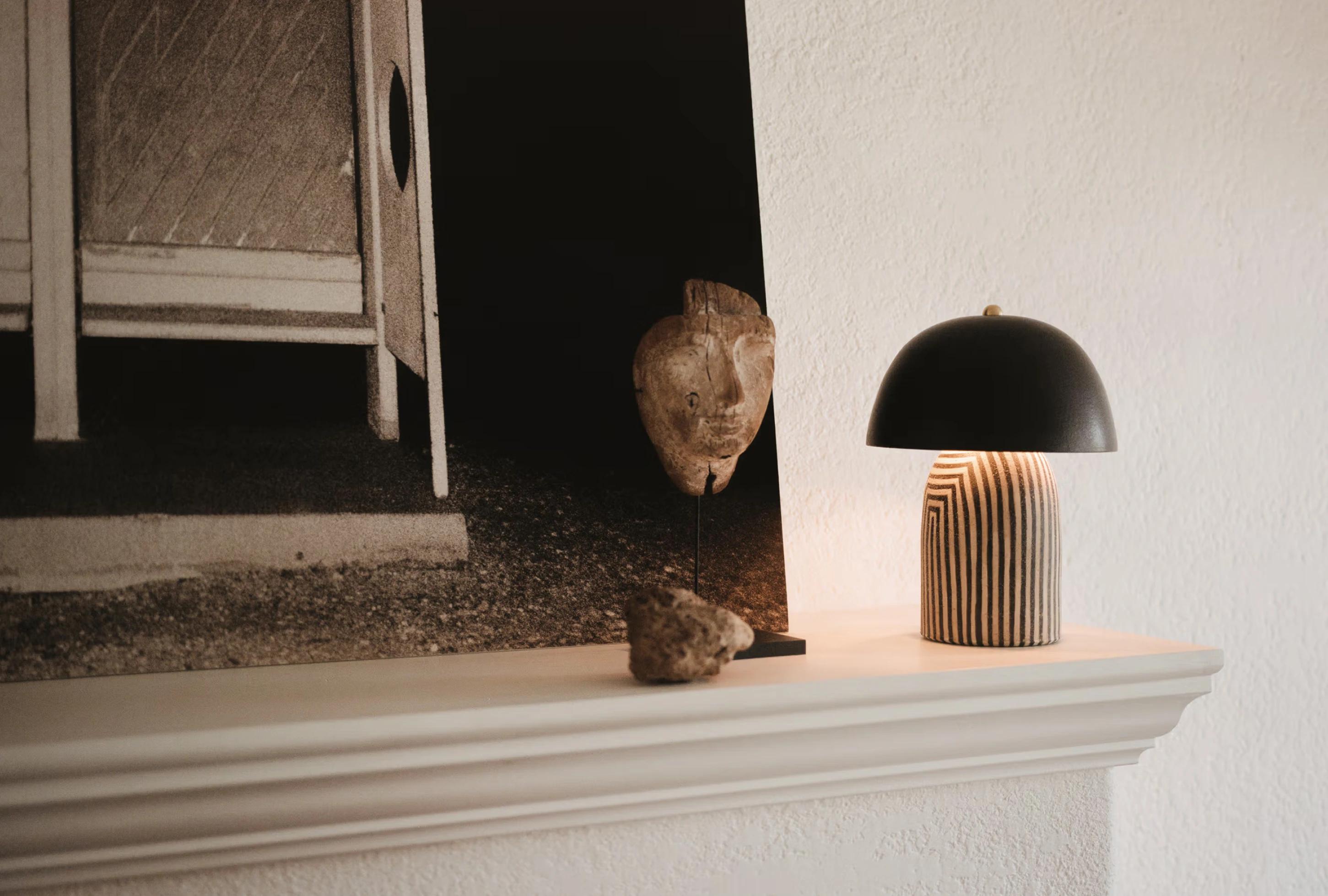 A wooden sculpture of a face, decorative stone, and a striped lamp on a light-colored mantel with a black and white photo ...