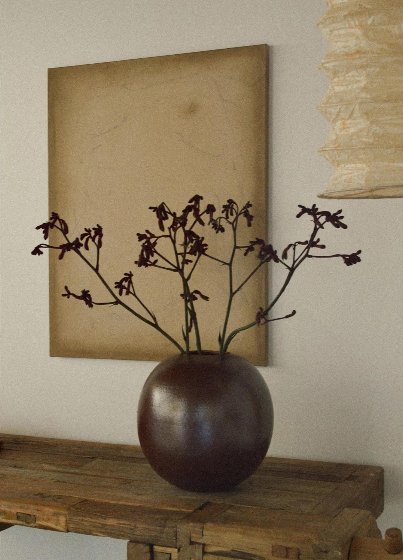 A round brown vase holds dark flowers, set against a beige backdrop and rustic wooden table.