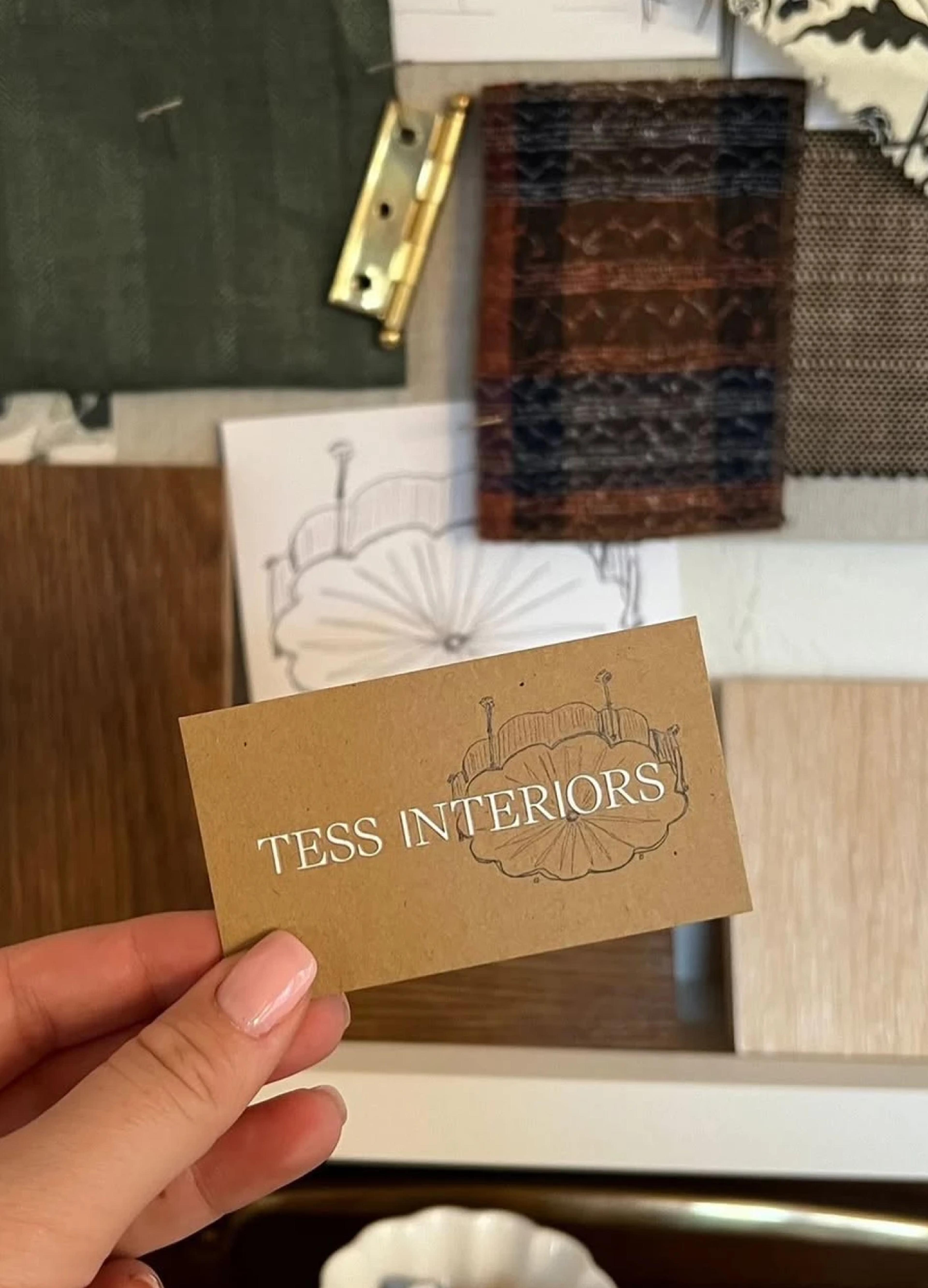 A hand holds a business card for "Tess Interiors" amidst fabric samples, sketches, and wood materials on a design board.