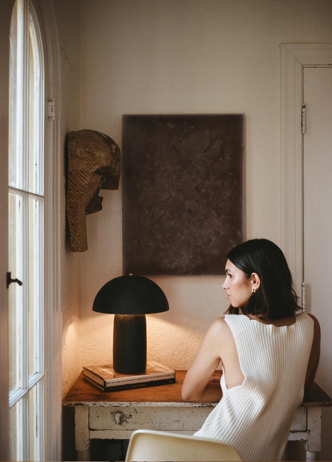 A woman in a sleeveless white top sits at a desk, illuminated by a stylish lamp and surrounded by art and decor.