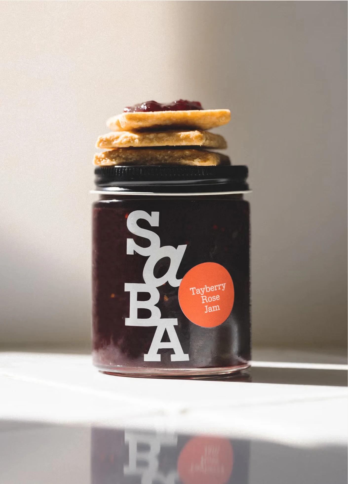 A jar of Tayberry Rose Jam topped with stacked cookies, set against a softly lit background.