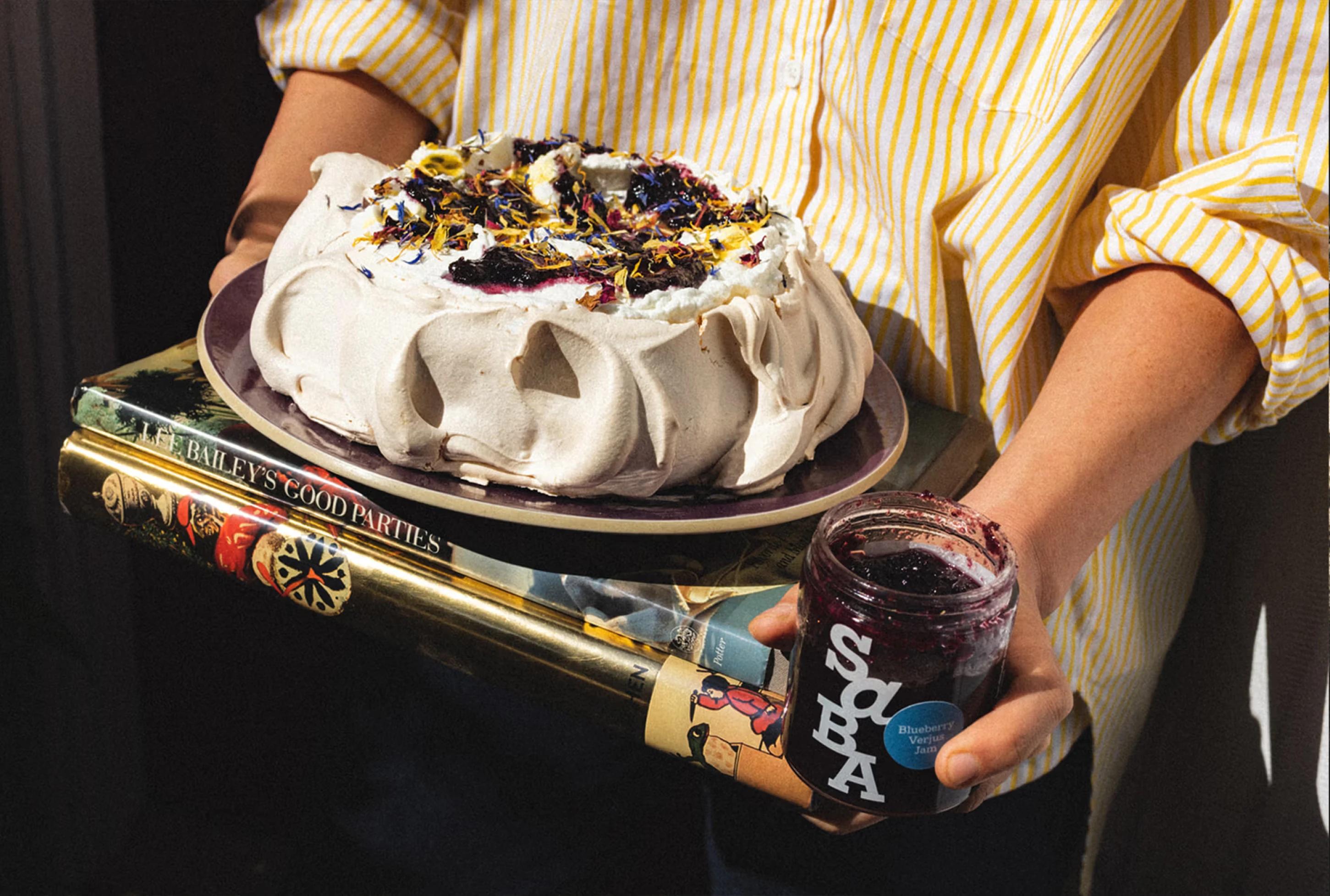 A person holds a pavlova cake on a plate, with a jar of blueberry jam and books stacked beneath it.