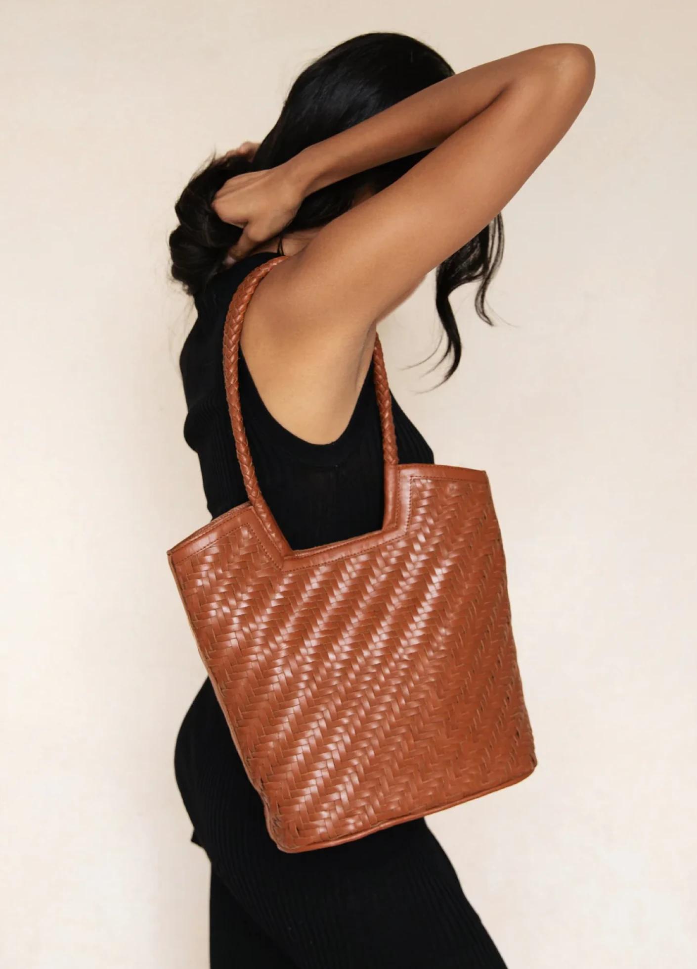A model in a black outfit gracefully holds a woven brown tote bag against a soft, neutral background.
