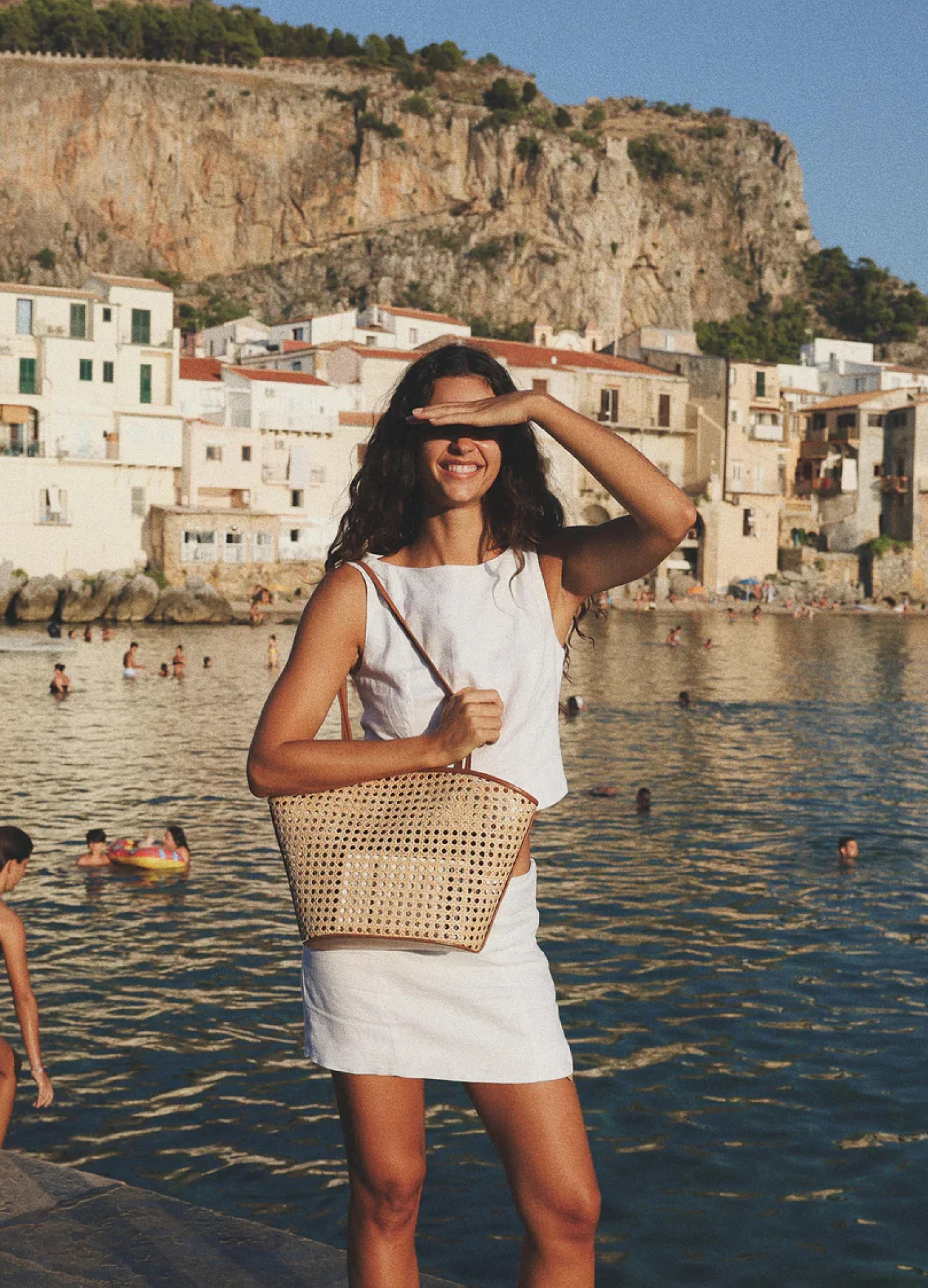A smiling woman in a white outfit stands by the water, shielding her eyes from the sun, with beachgoers in the background.