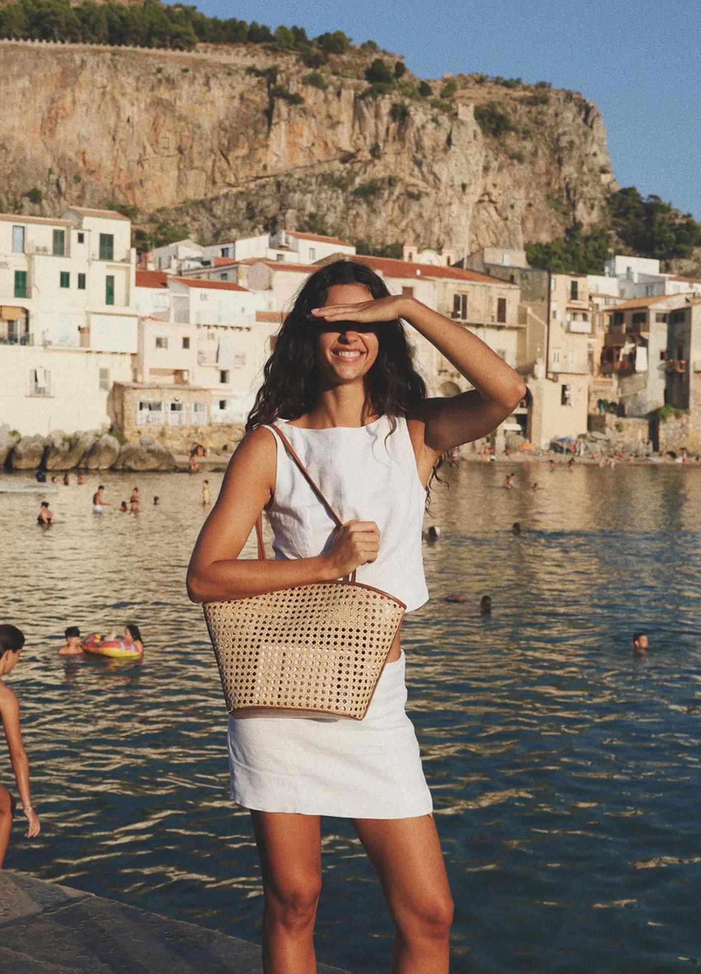A smiling woman in a white outfit stands by the water, shielding her eyes from the sun, with beachgoers in the background.