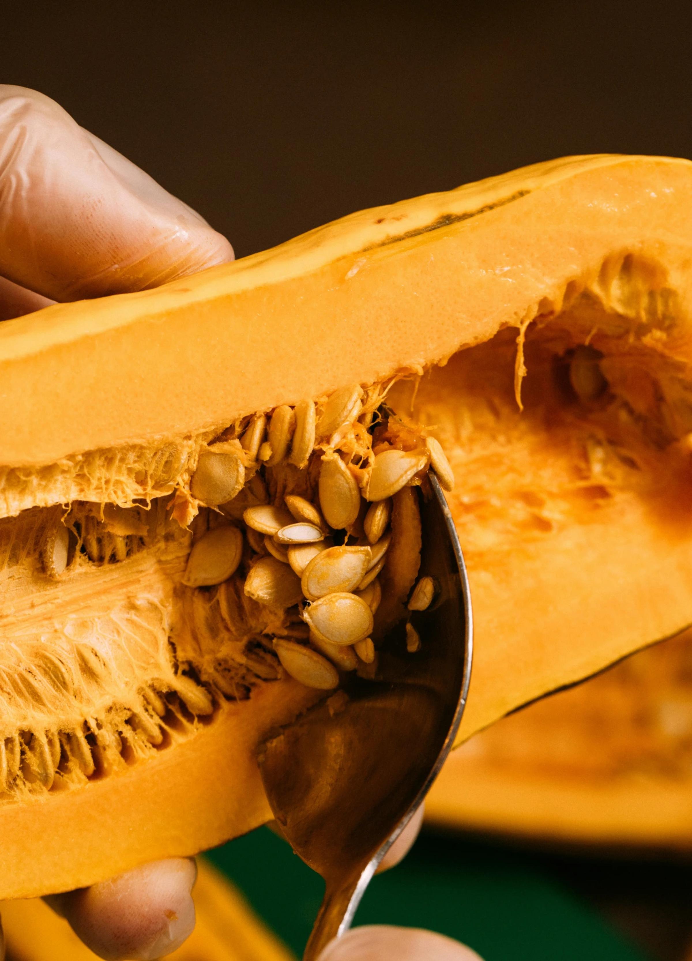 A hand holding a halved butternut squash, revealing seeds and fibrous strands inside. A spoon is scooping out the seeds.