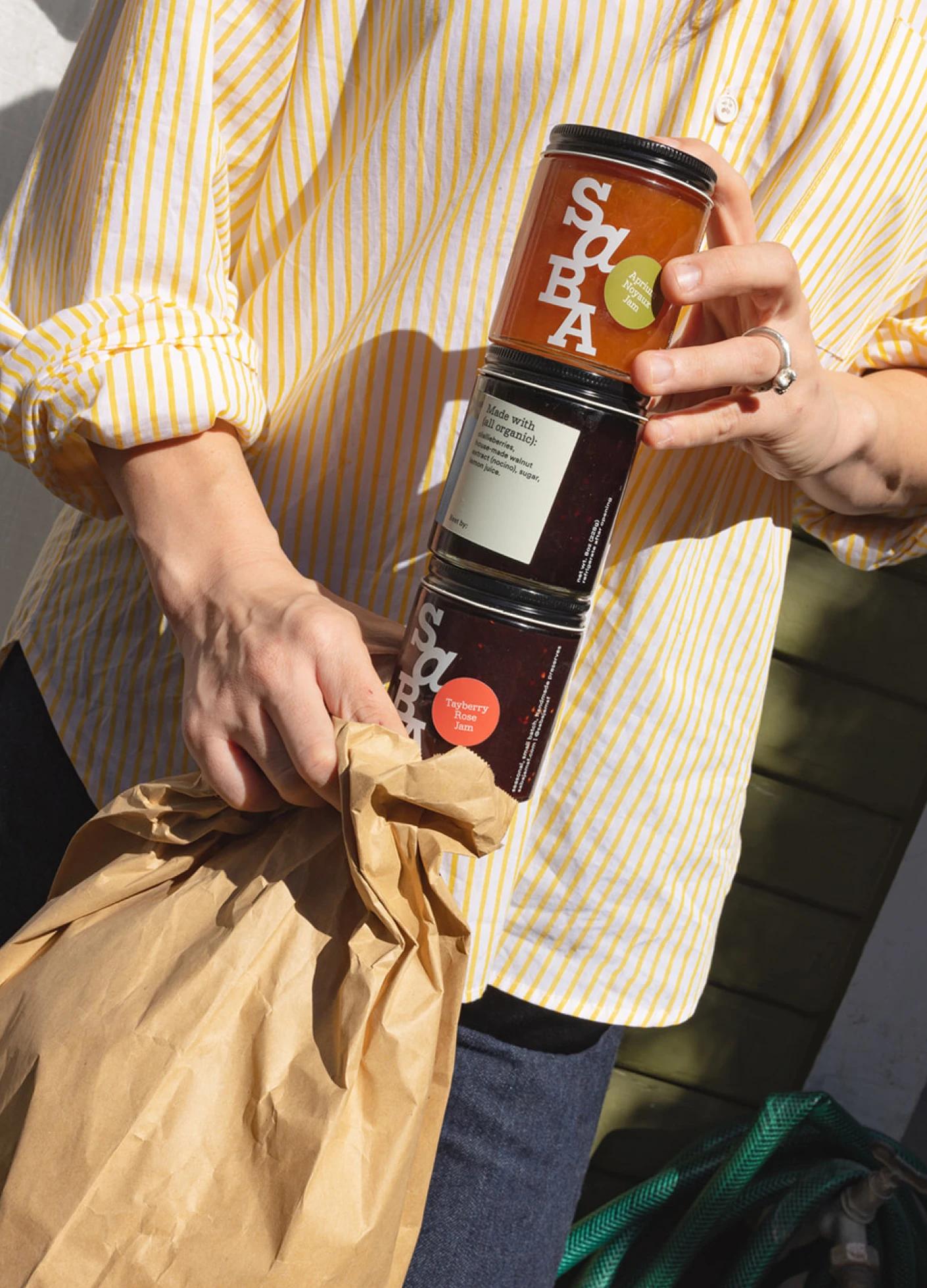 A person in a striped yellow shirt holds three jars of jam and a brown paper bag.