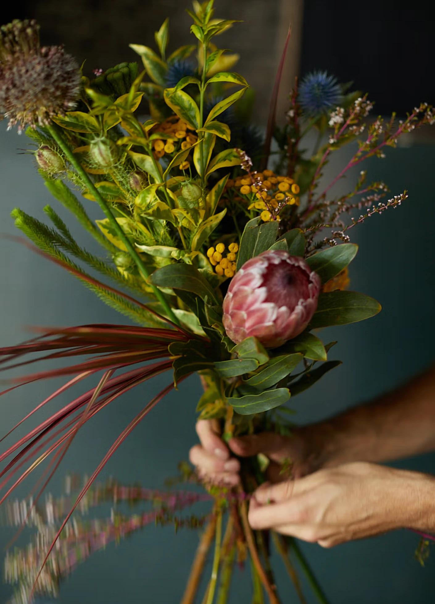 A person arranging a colorful bouquet of diverse flowers and greenery against a muted background.