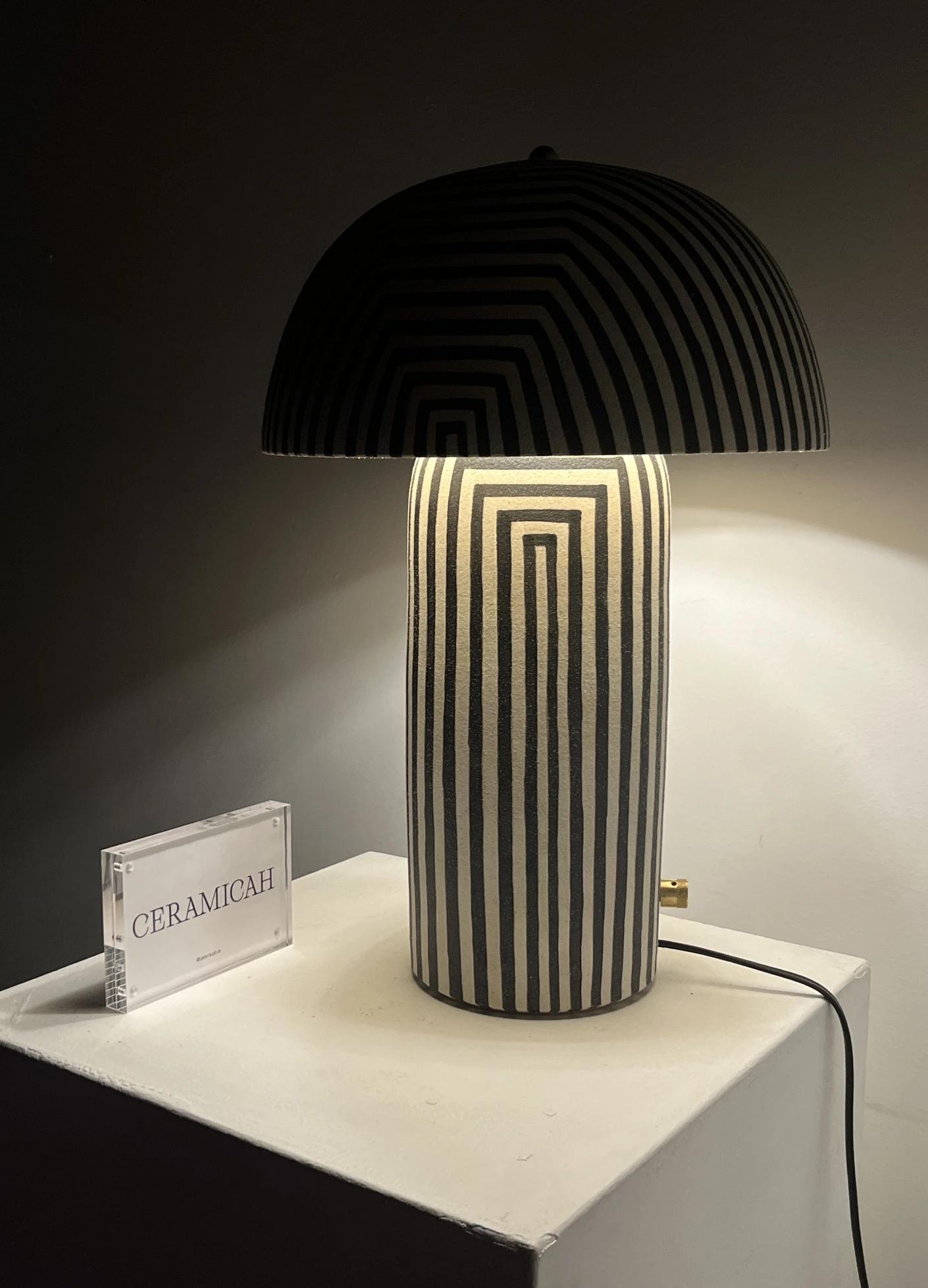 A stylish striped lamp with a rounded shade sits on a pedestal beside a clear sign displaying "CERAMICAH."