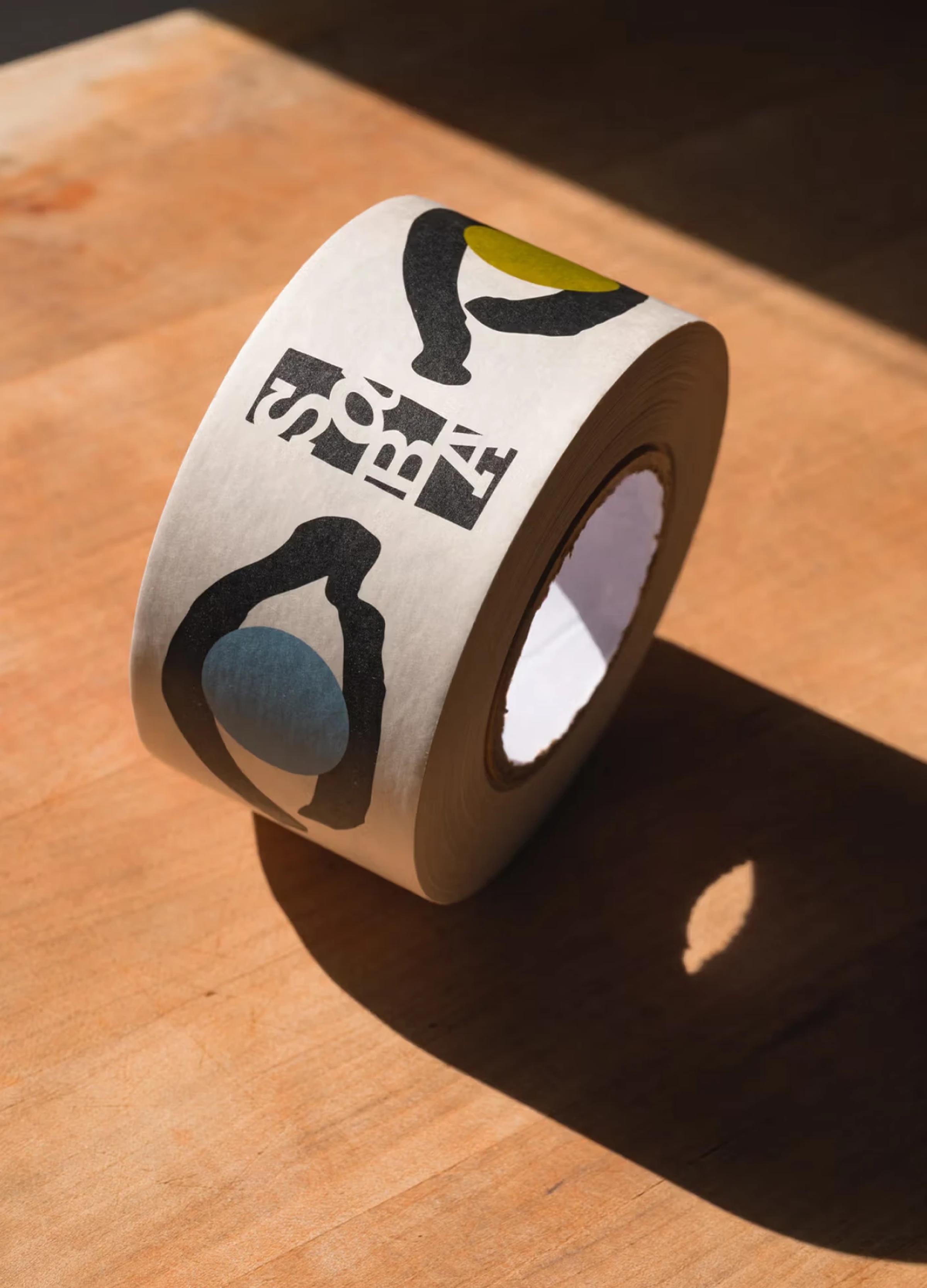 A roll of decorative tape with colorful abstract shapes and letters on a wooden surface, casting a shadow.