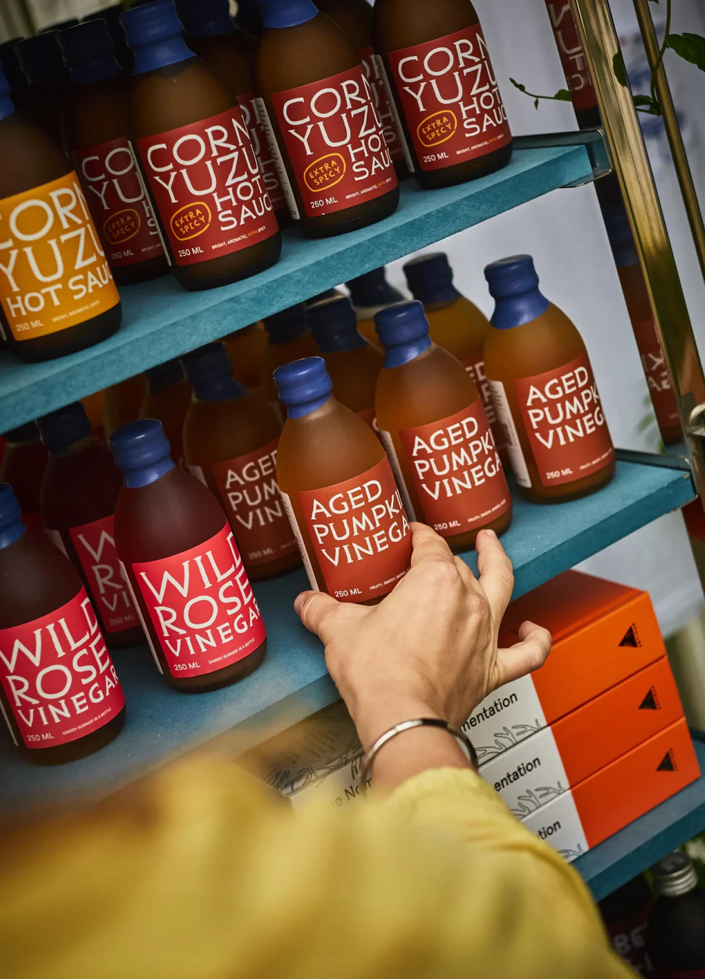 Glass bottles of various sauces and vinegars displayed on teal shelves, with a hand reaching for a bottle.