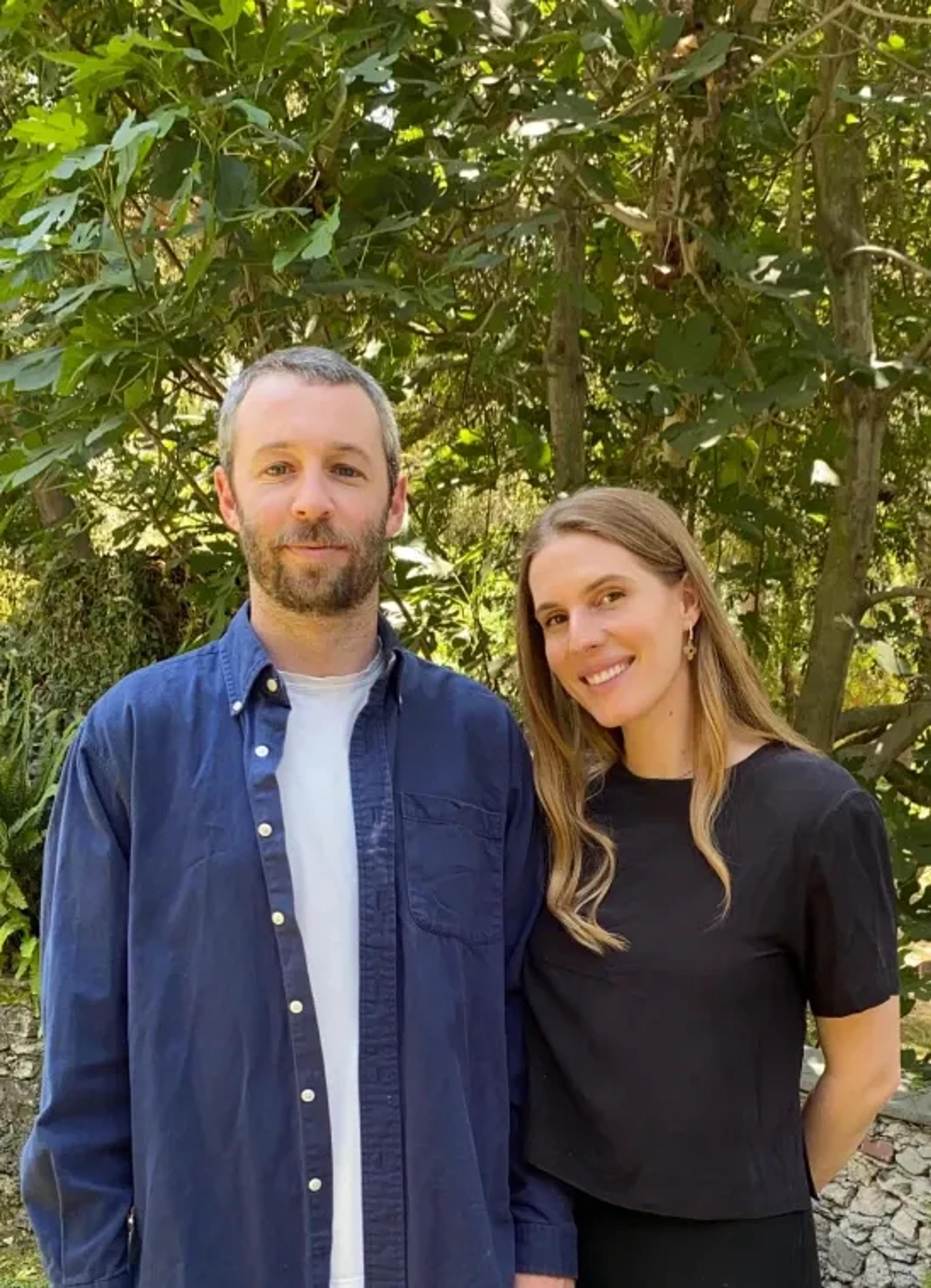 A man with a beard and a woman with long hair stand together, smiling, surrounded by greenery.