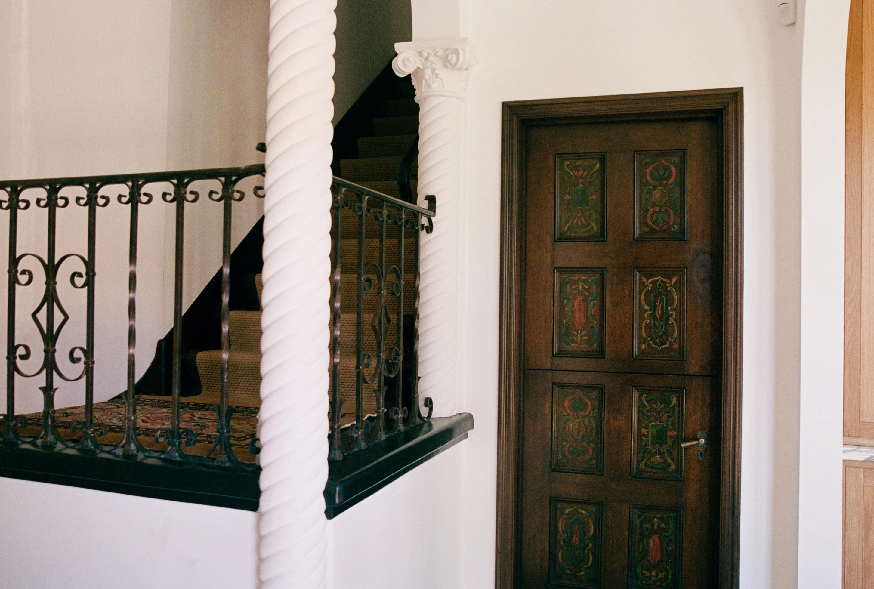 A staircase with decorative iron railing leads to an ornate wooden door, featuring colorful carvings, in a bright hallway.